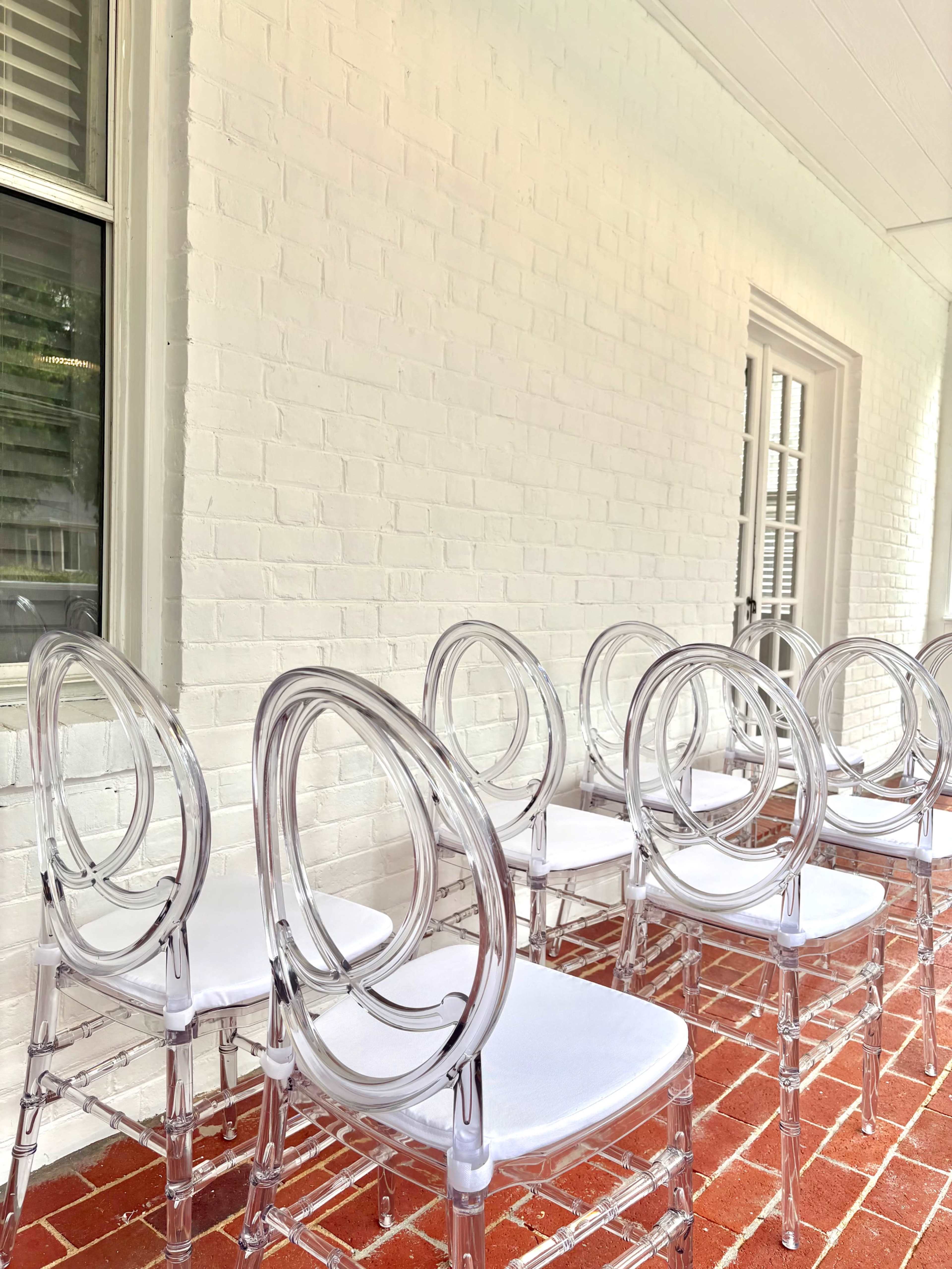 A row of clear acrylic chairs with white cushions is positioned against a white brick wall.