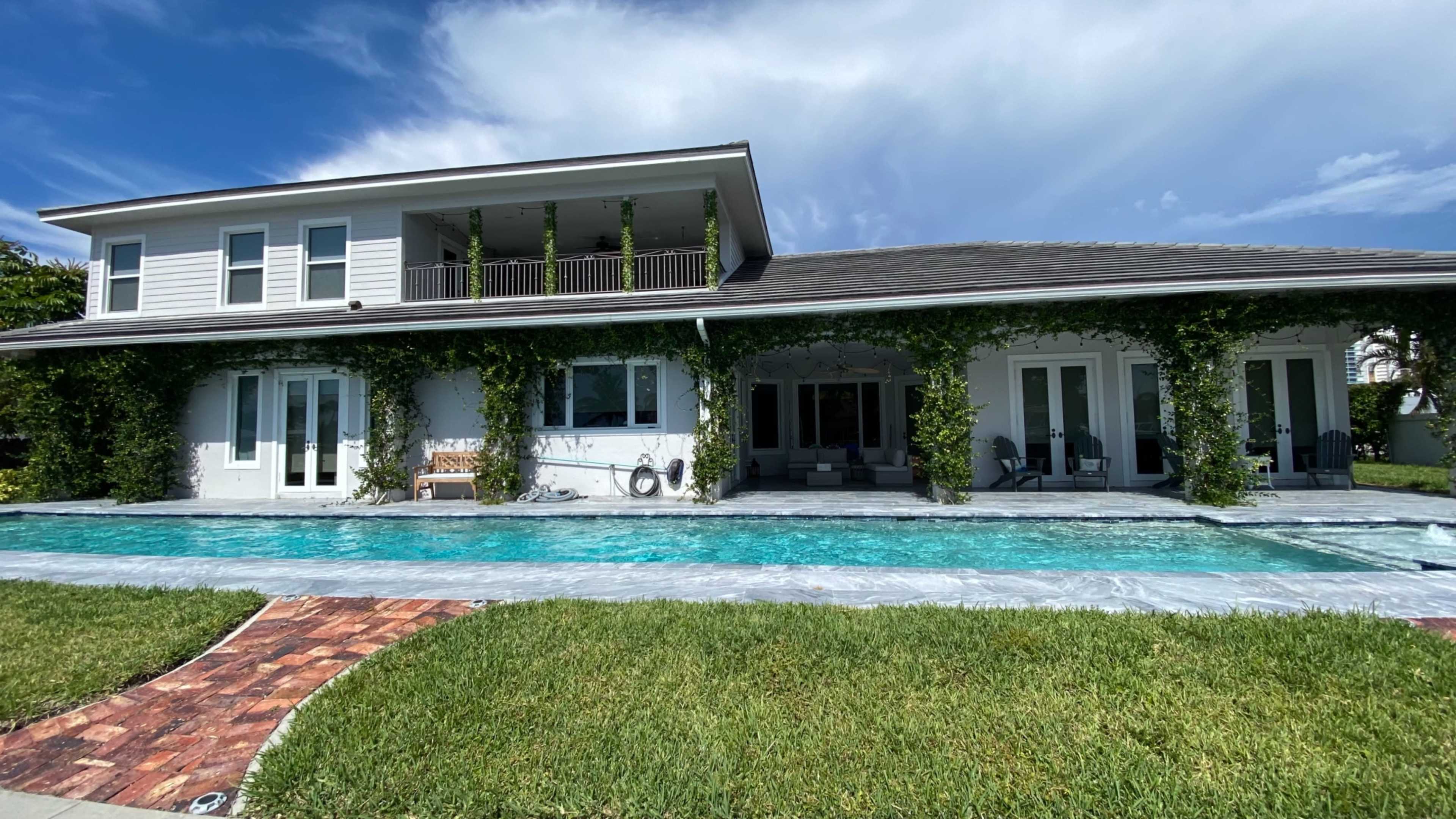 A two-story house with a pool in the foreground is surrounded by green grass and climbing vines on the walls.