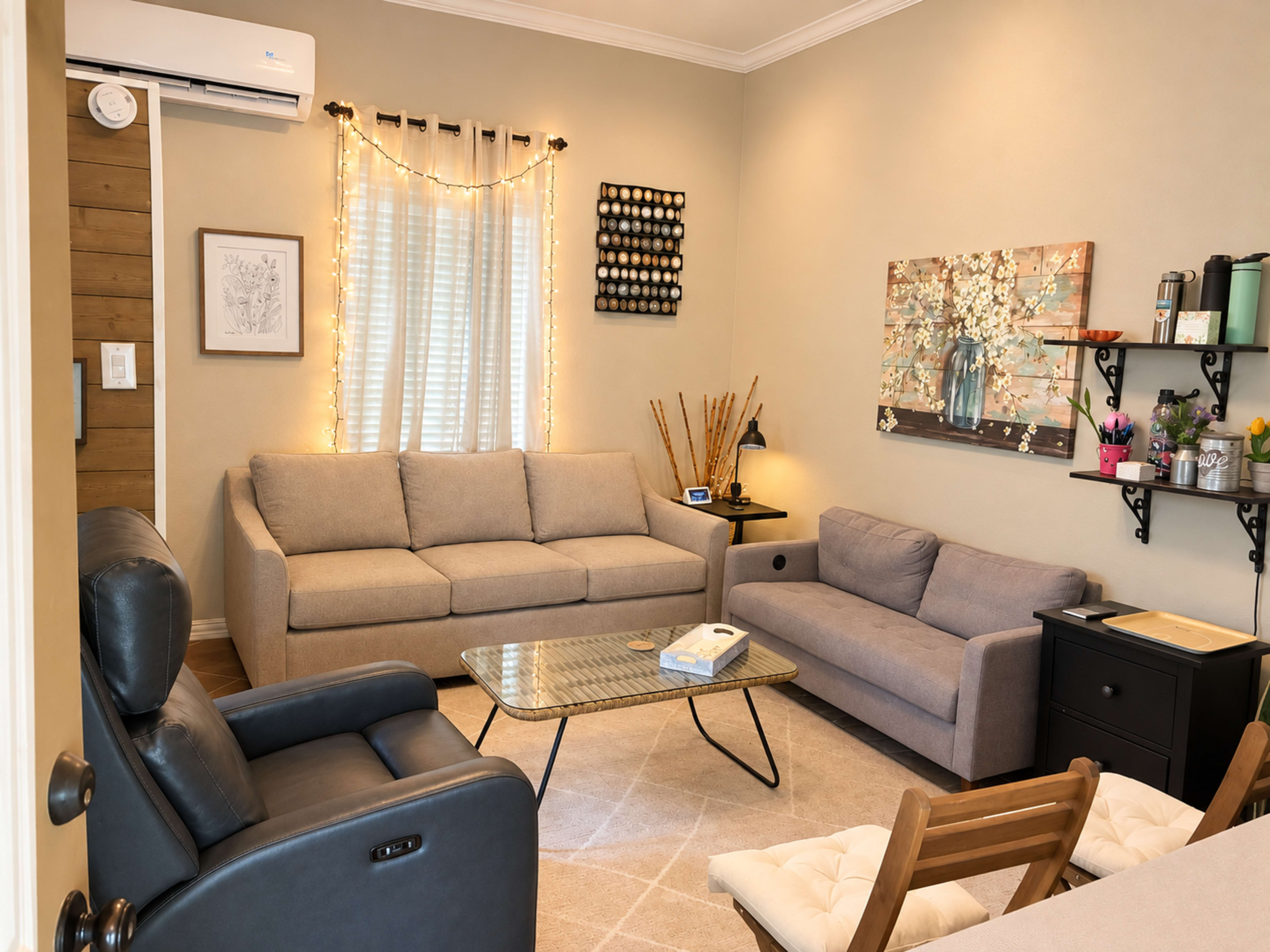 The image shows a cozy living room with a gray sofa, a smaller gray couch, a black chair, and a glass coffee table, all arranged around a light-colored carpet.