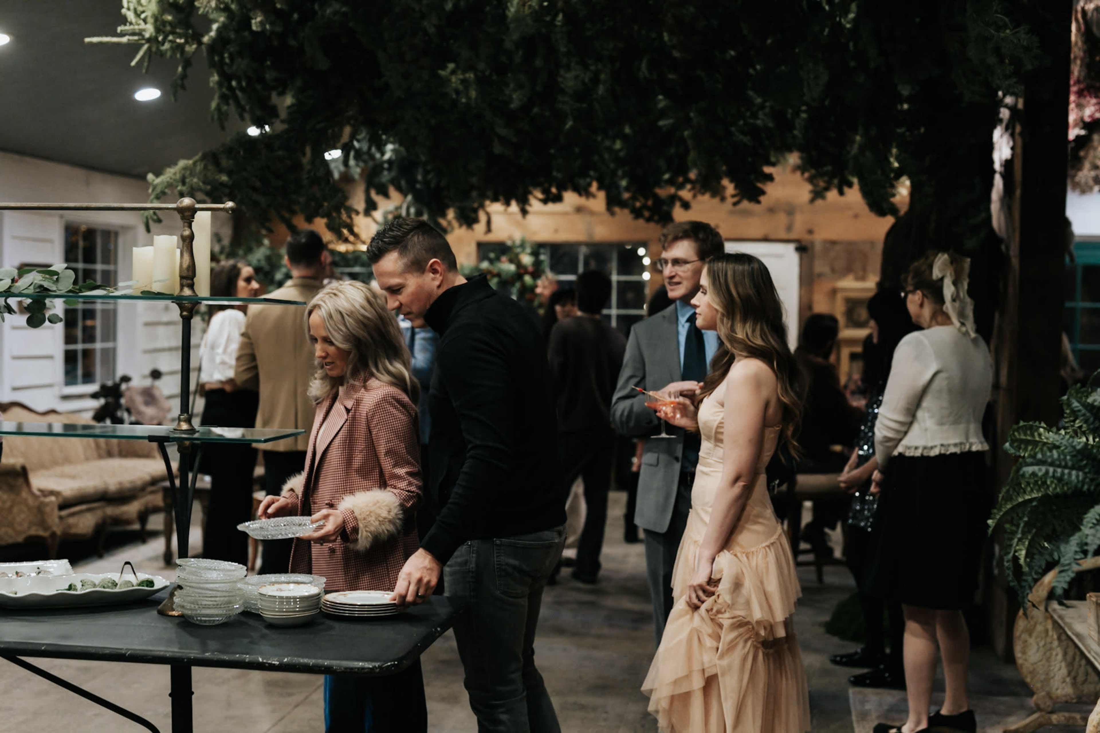 A group of people socializes at a gathering, with some individuals standing by a table that has plates and food, while others chat in the background.