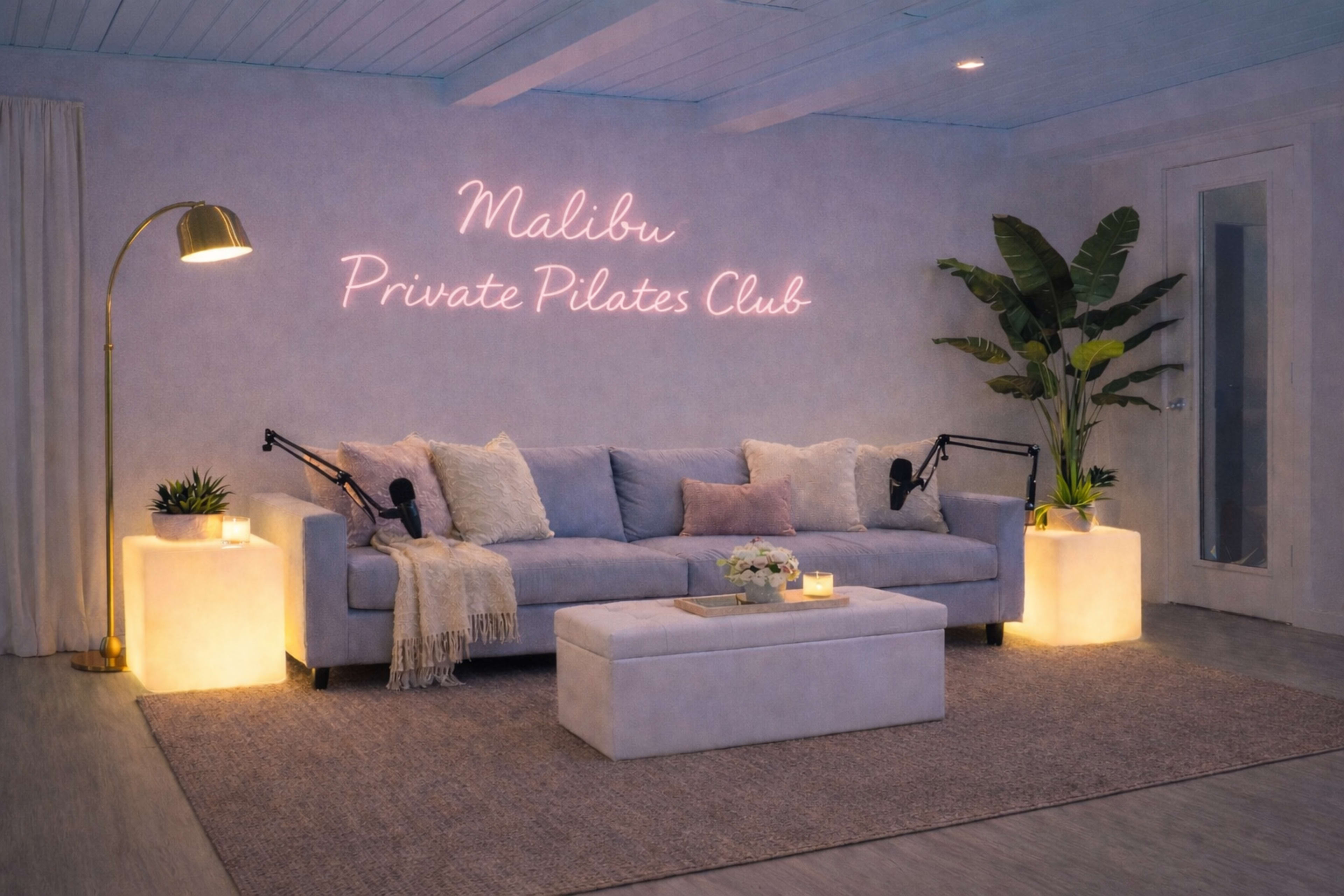 The image shows a modern, well-lit room with a soft gray couch, decorative plants, and illuminated pedestals, featuring a neon sign that reads "Malibu Private Pilates Club" on the wall.