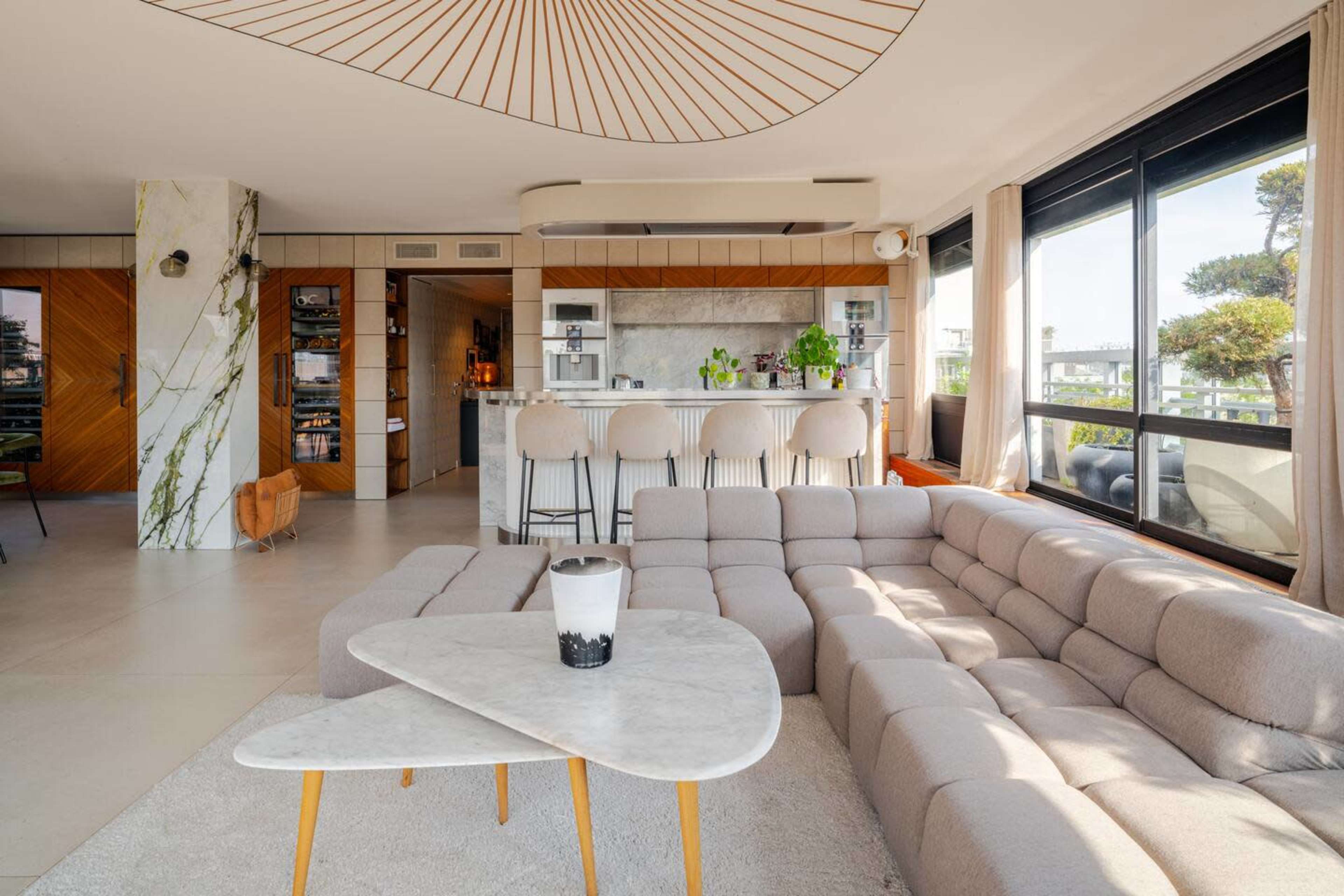 A modern living room with a large, light-colored sectional sofa, a marble coffee table, and a bar area in the background featuring greenery and stylish stools.