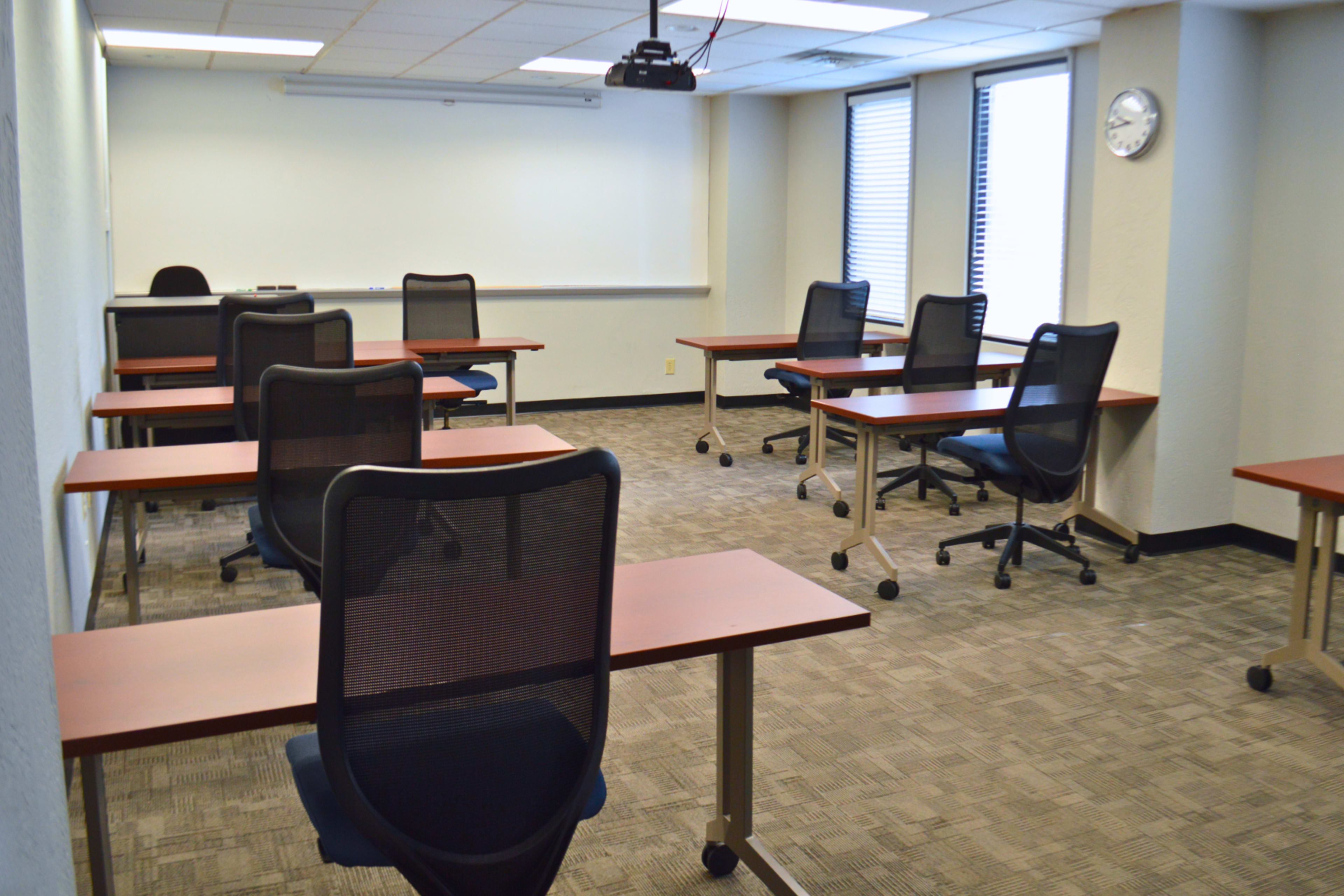 A classroom with several empty desks and chairs arranged in a spacious layout, featuring a whiteboard and a clock on the wall.