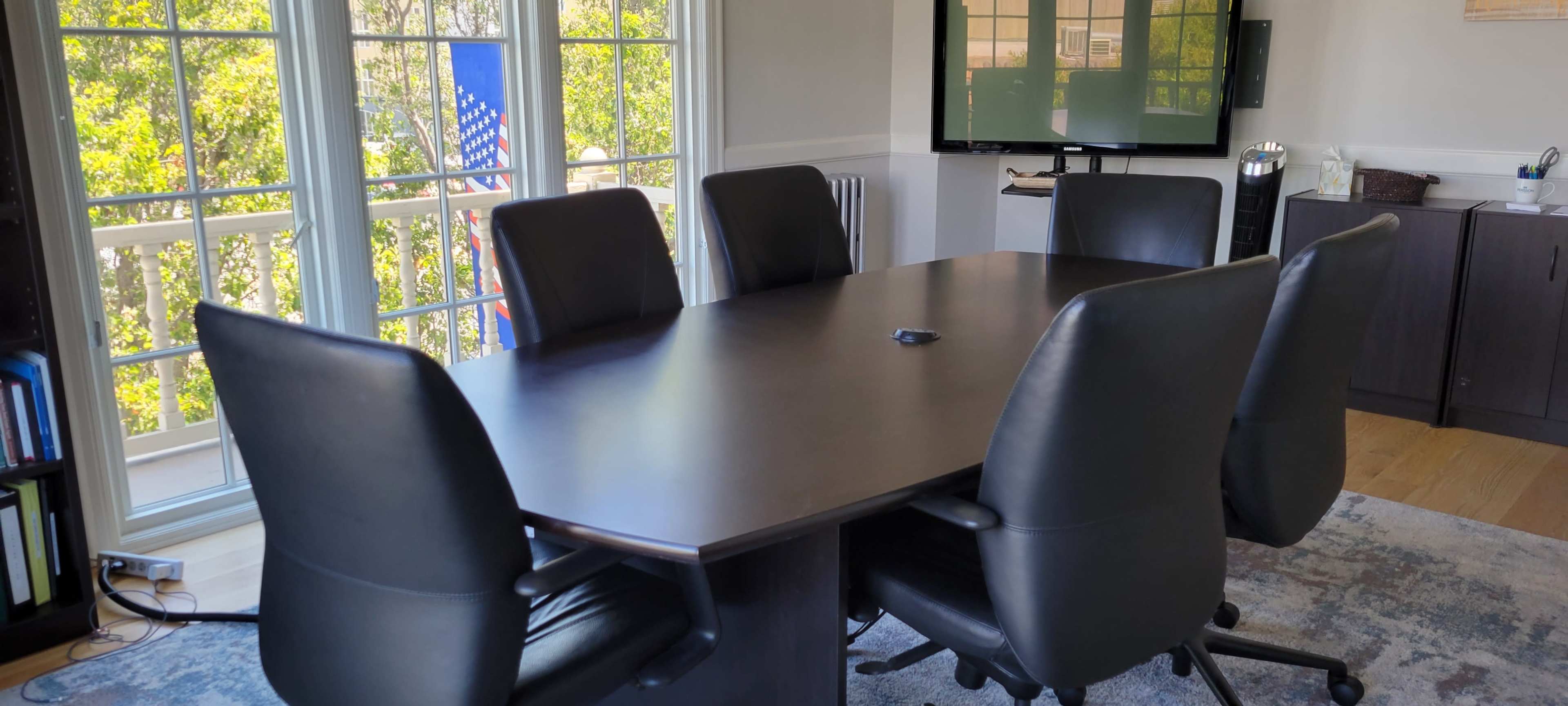 A modern conference room features a large table surrounded by six black ergonomic chairs, with windows displaying an American flag in the background.