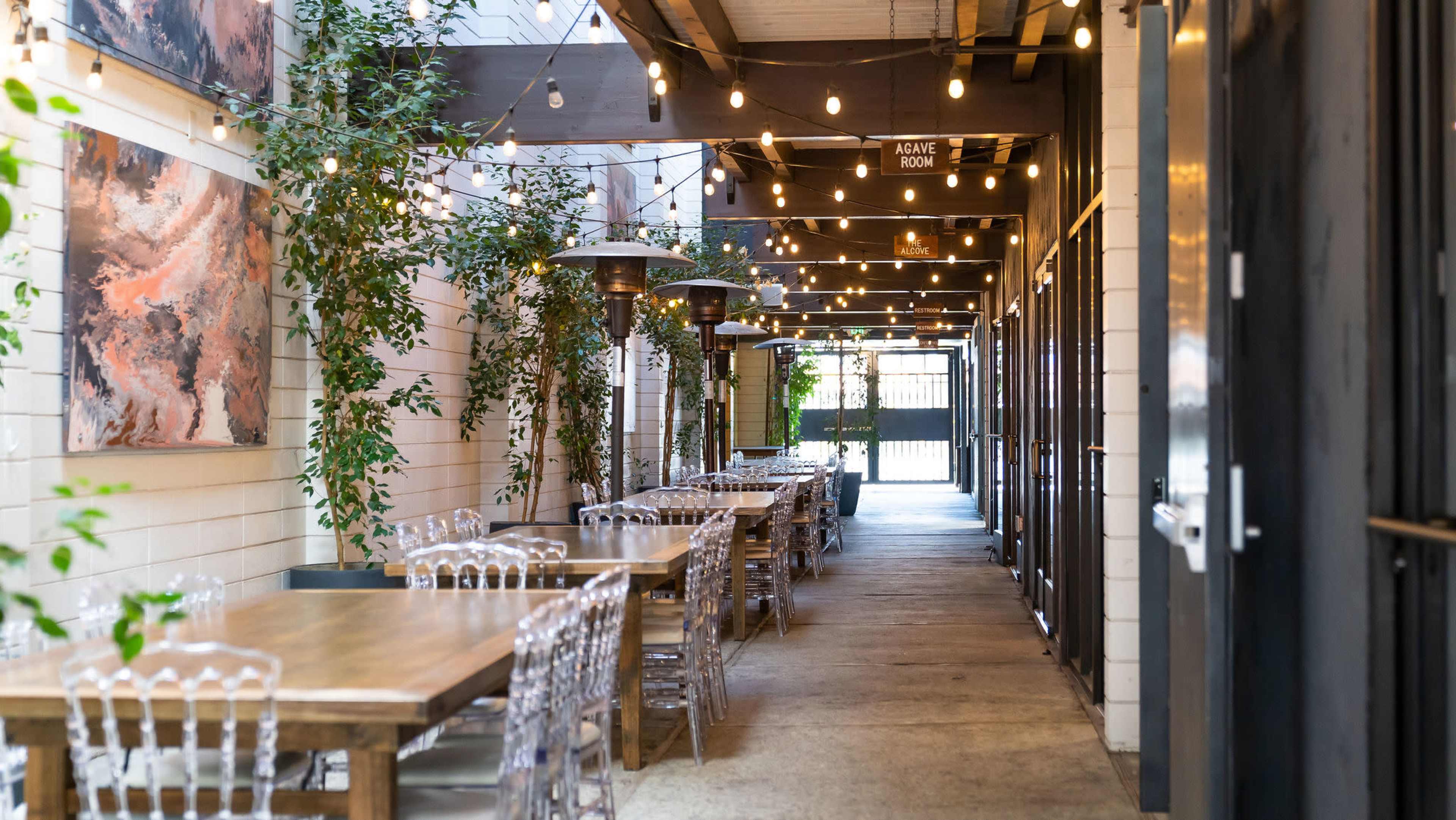A narrow outdoor walkway lined with dining tables and string lights, flanked by plants and open doorways.