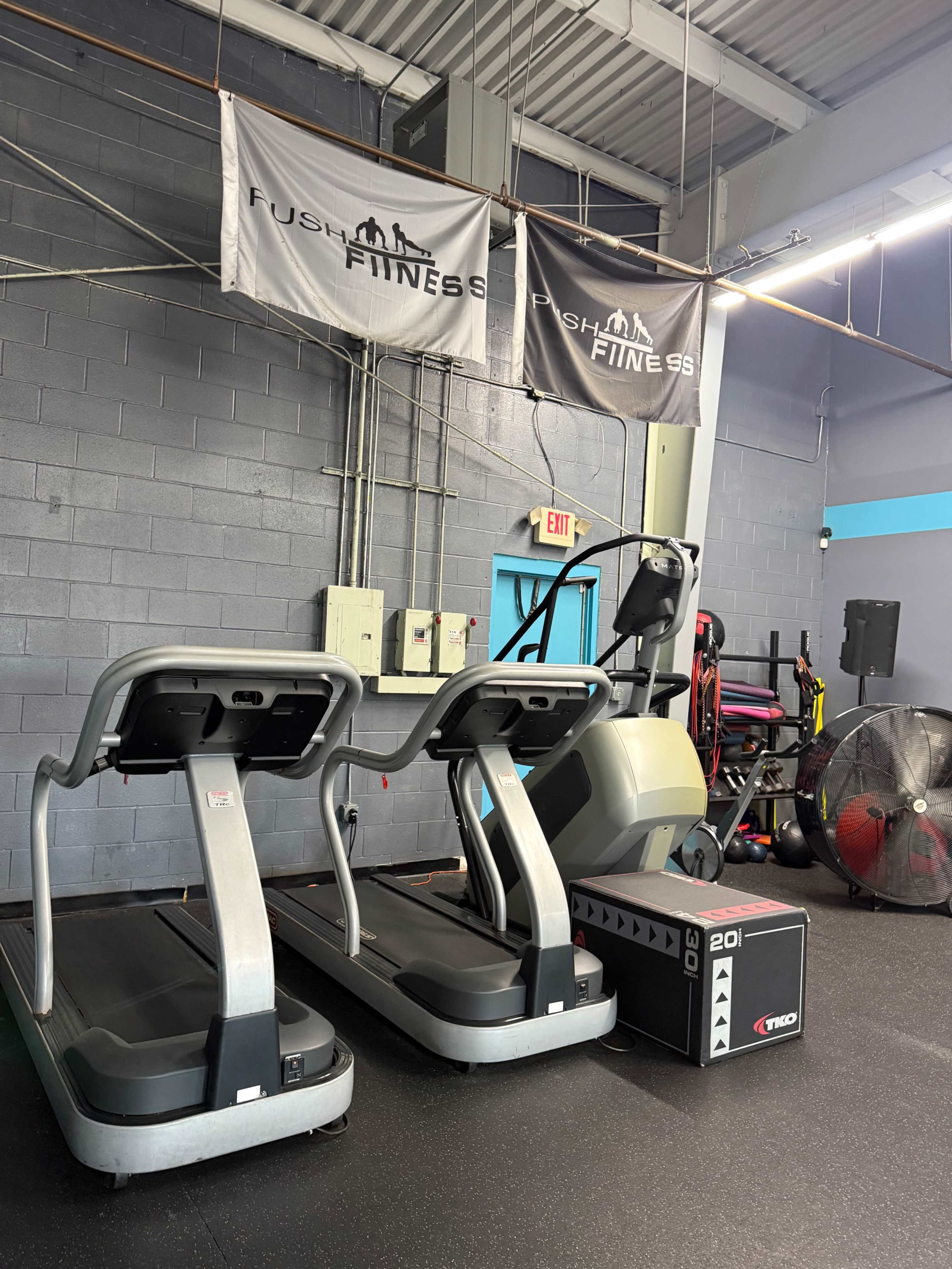 The image shows two treadmills and an elliptical machine in a gym, with fitness banners hanging on the wall and various exercise equipment in the background.