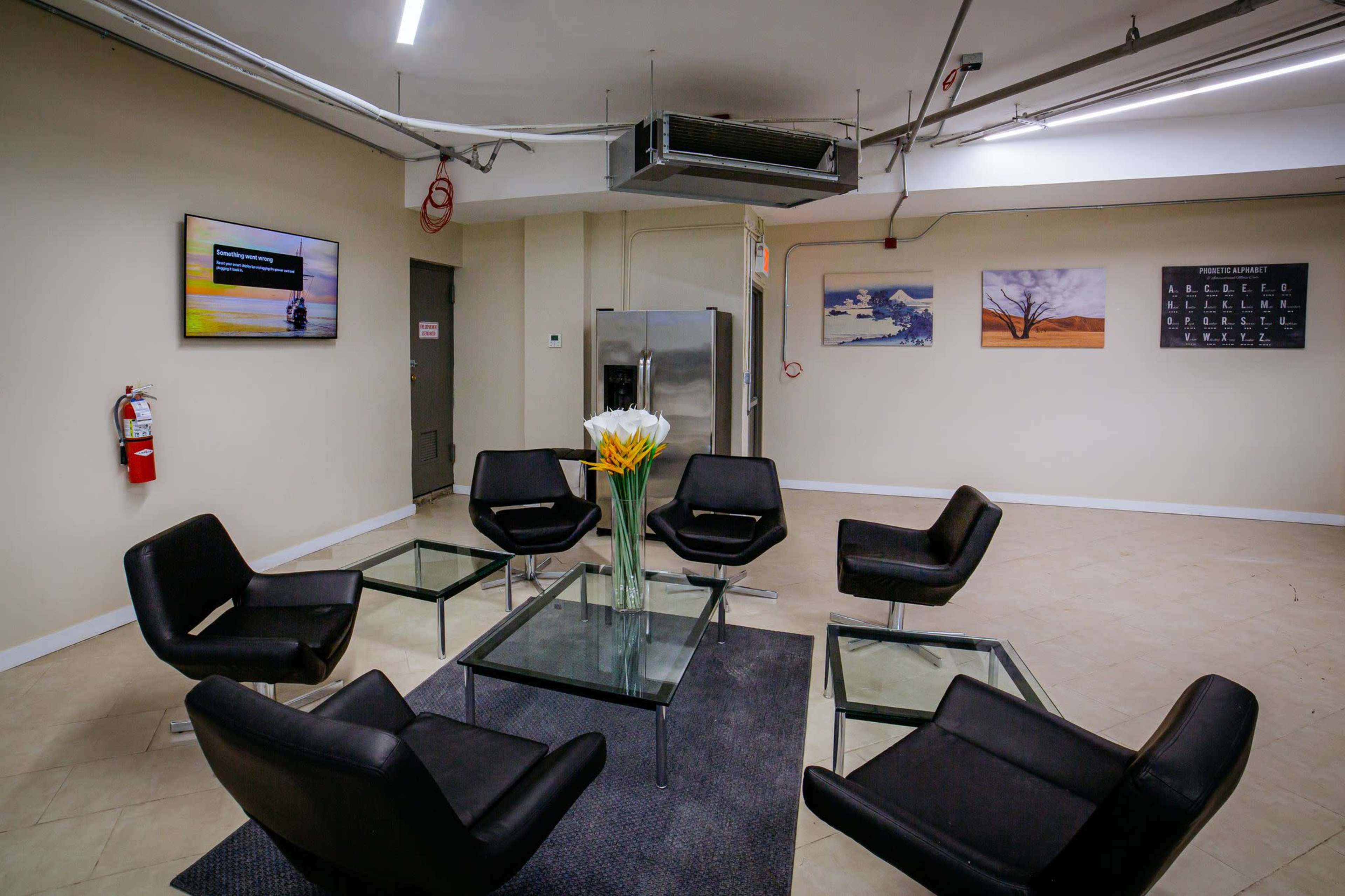 A waiting area features six black chairs arranged around a glass table, with a flower arrangement on top, and a television mounted on the wall.