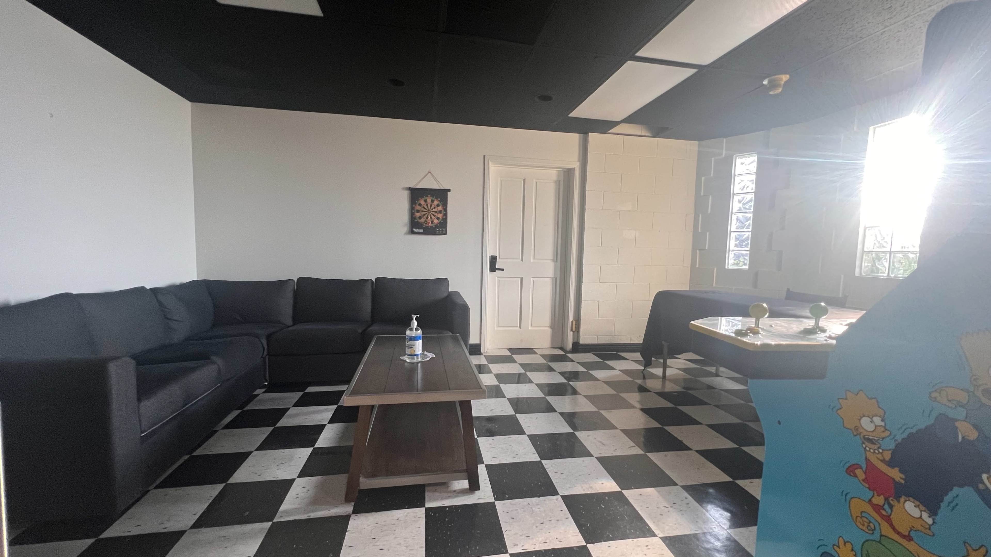 A lounge area with a black and white checkered floor, a dark couch, a coffee table, and a dartboard on the wall.