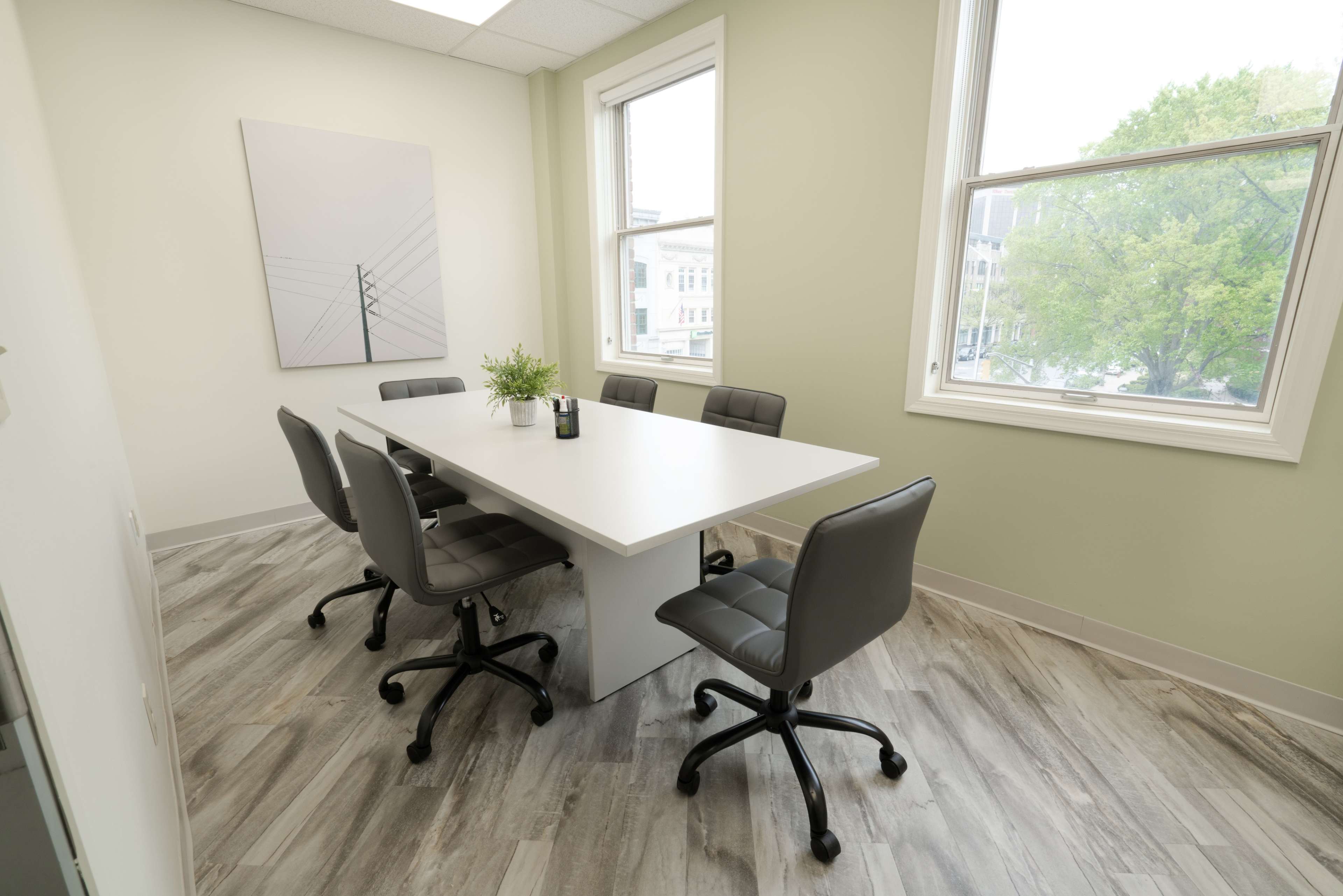 A modern conference room with a large white table surrounded by six gray office chairs, featuring windows that allow natural light and a minimalist decor.