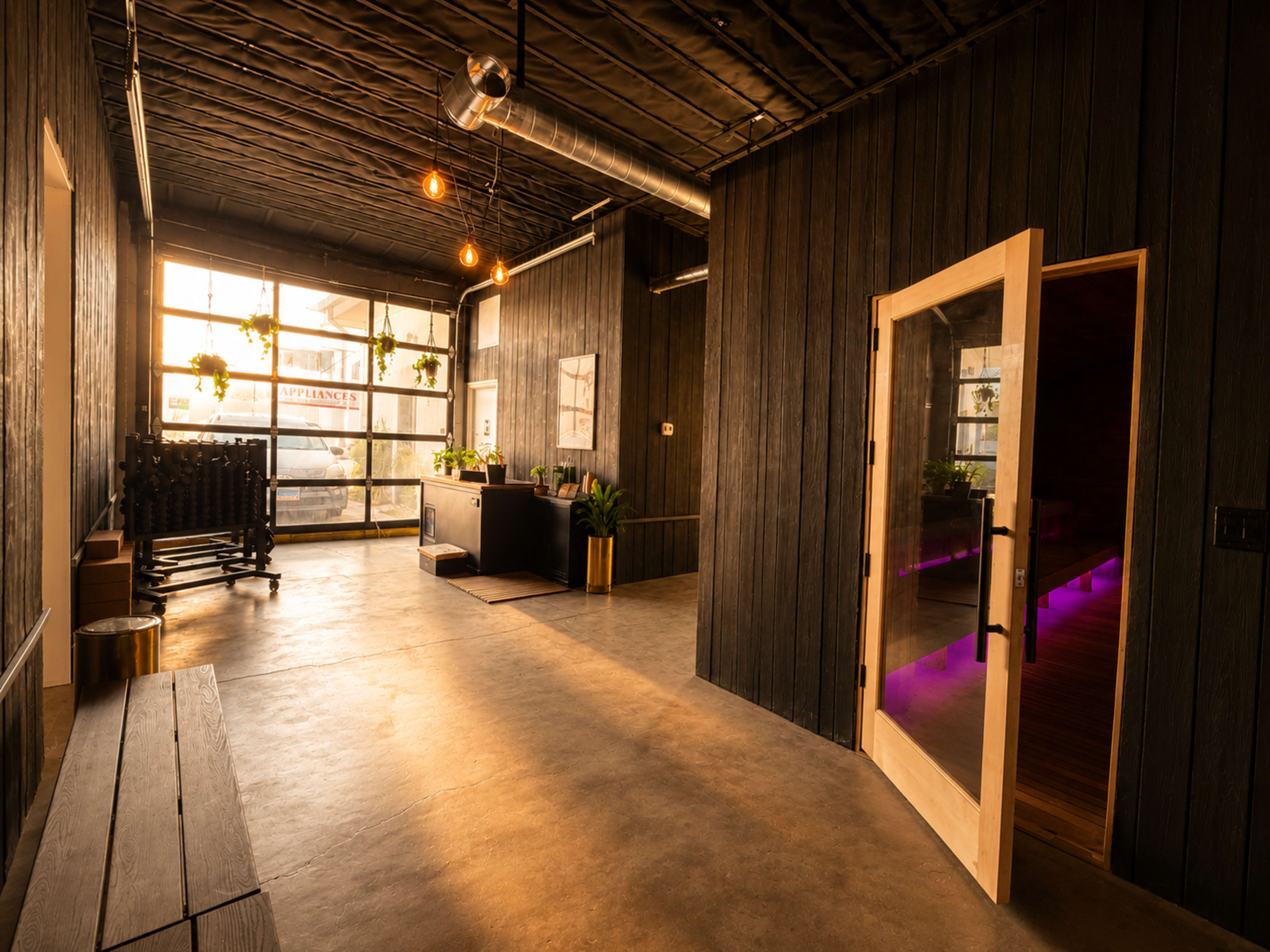 The image shows a modern interior space with wooden walls, large windows allowing natural light, and a reception area featuring plants and stylish decor.