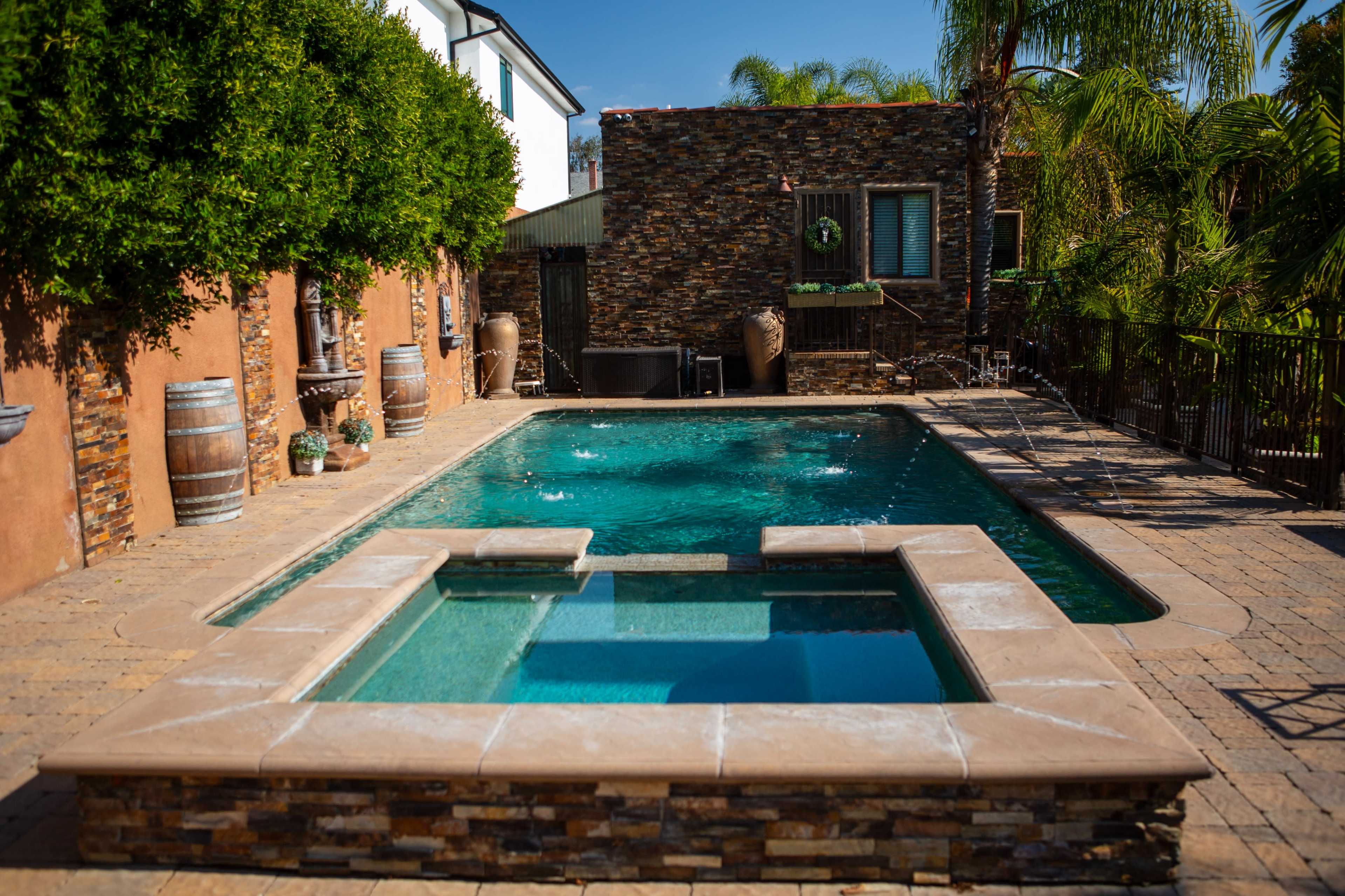 The image shows a swimming pool with an attached hot tub surrounded by stone walls, palm trees, and decorative barrels.