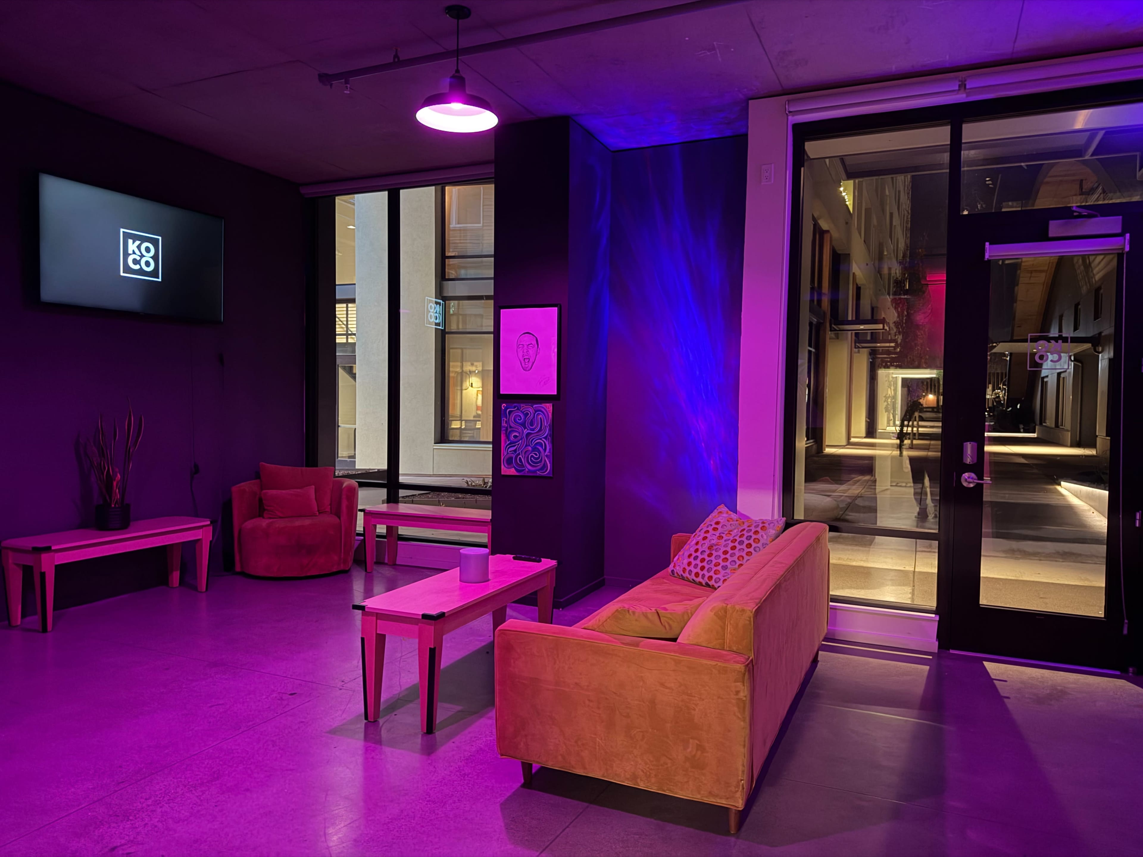The image shows a modern lounge area with purple lighting, featuring a sofa, armchairs, a coffee table, and large windows facing an outdoor walkway.