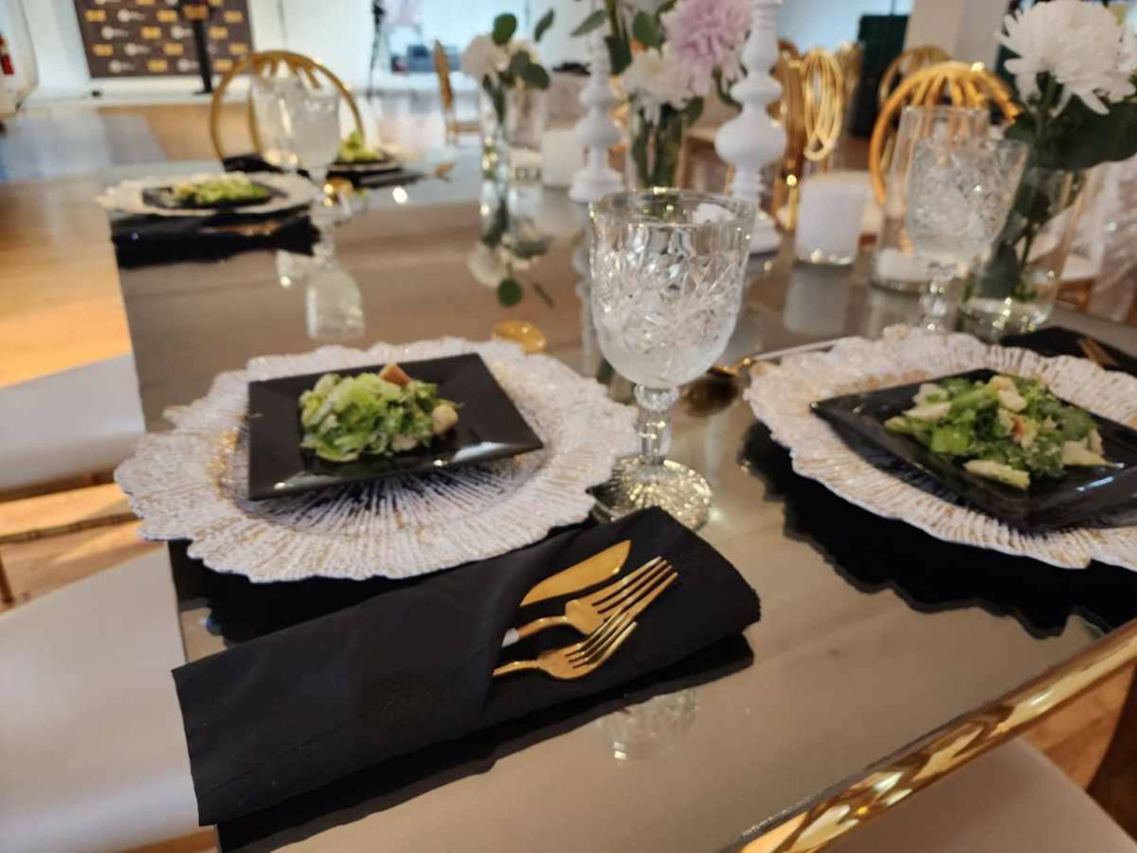 A dining table is set with black plates, crystal glasses, and elegant cutlery, surrounded by decorative flowers and a mirrored surface.