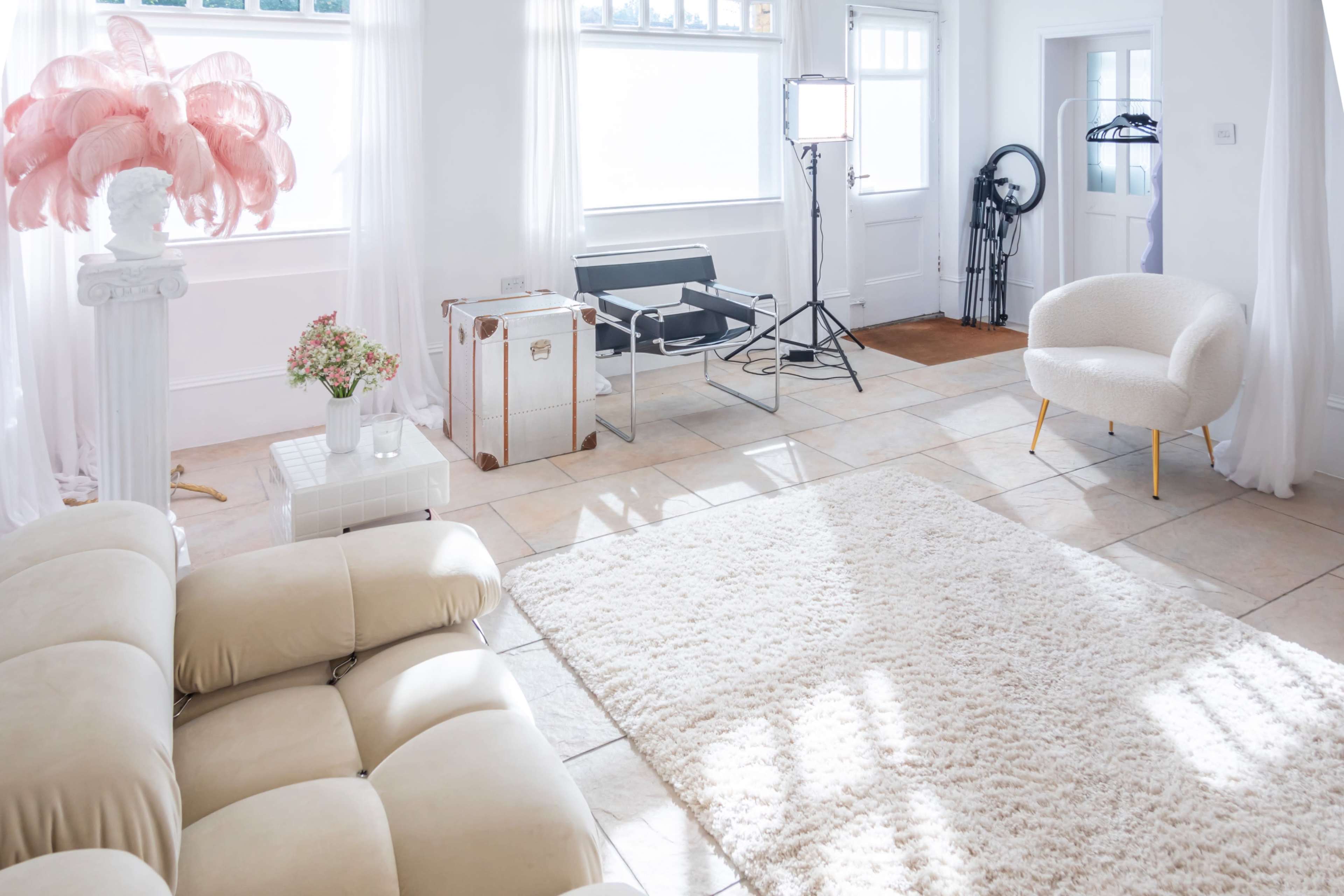 A bright, airy room with a plush area rug, a white sofa, a standing light setup, and a floral arrangement on a small table.