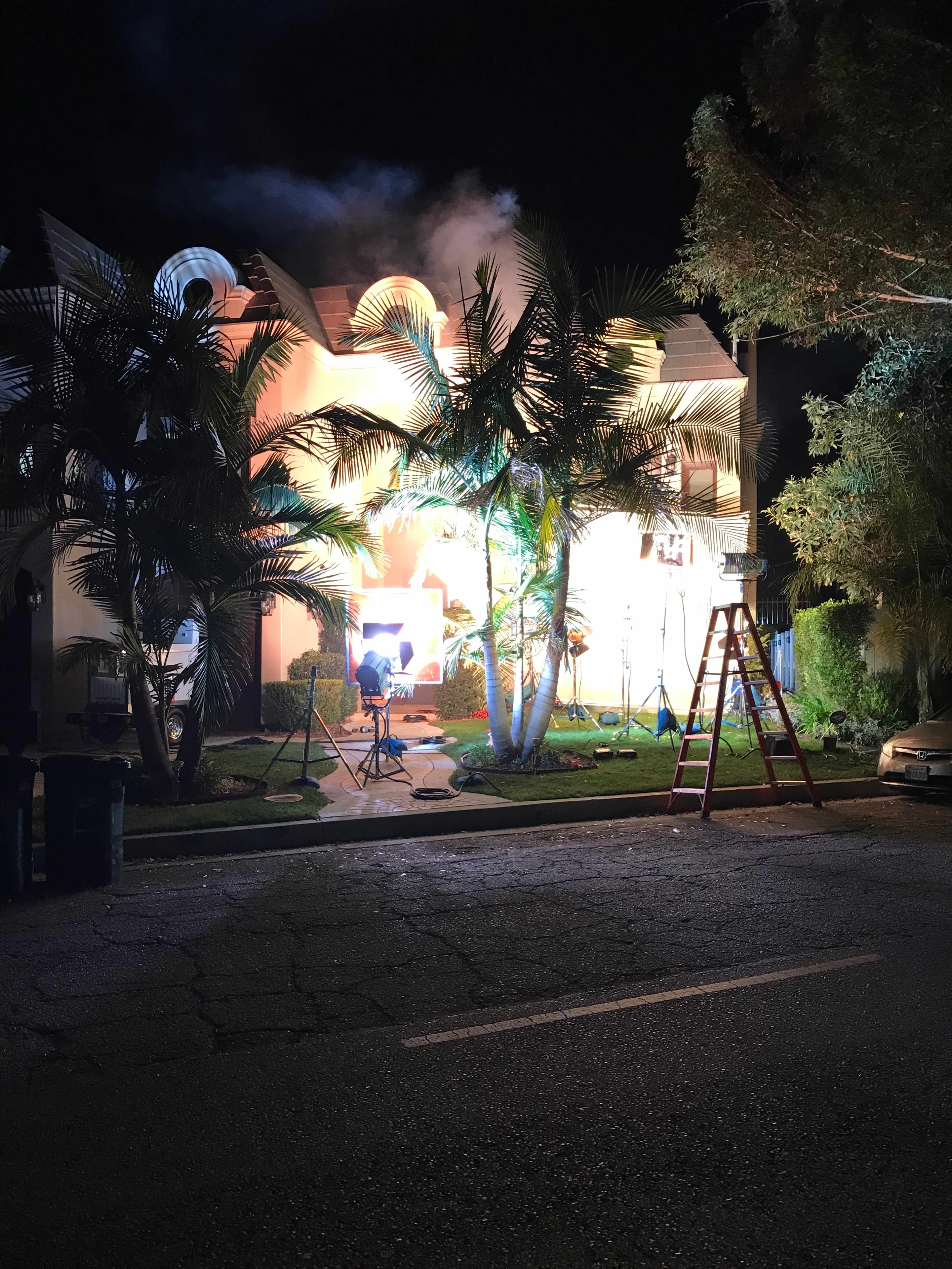 A film crew sets up equipment outside a house illuminated by bright lights while smoke billows from the windows at night.