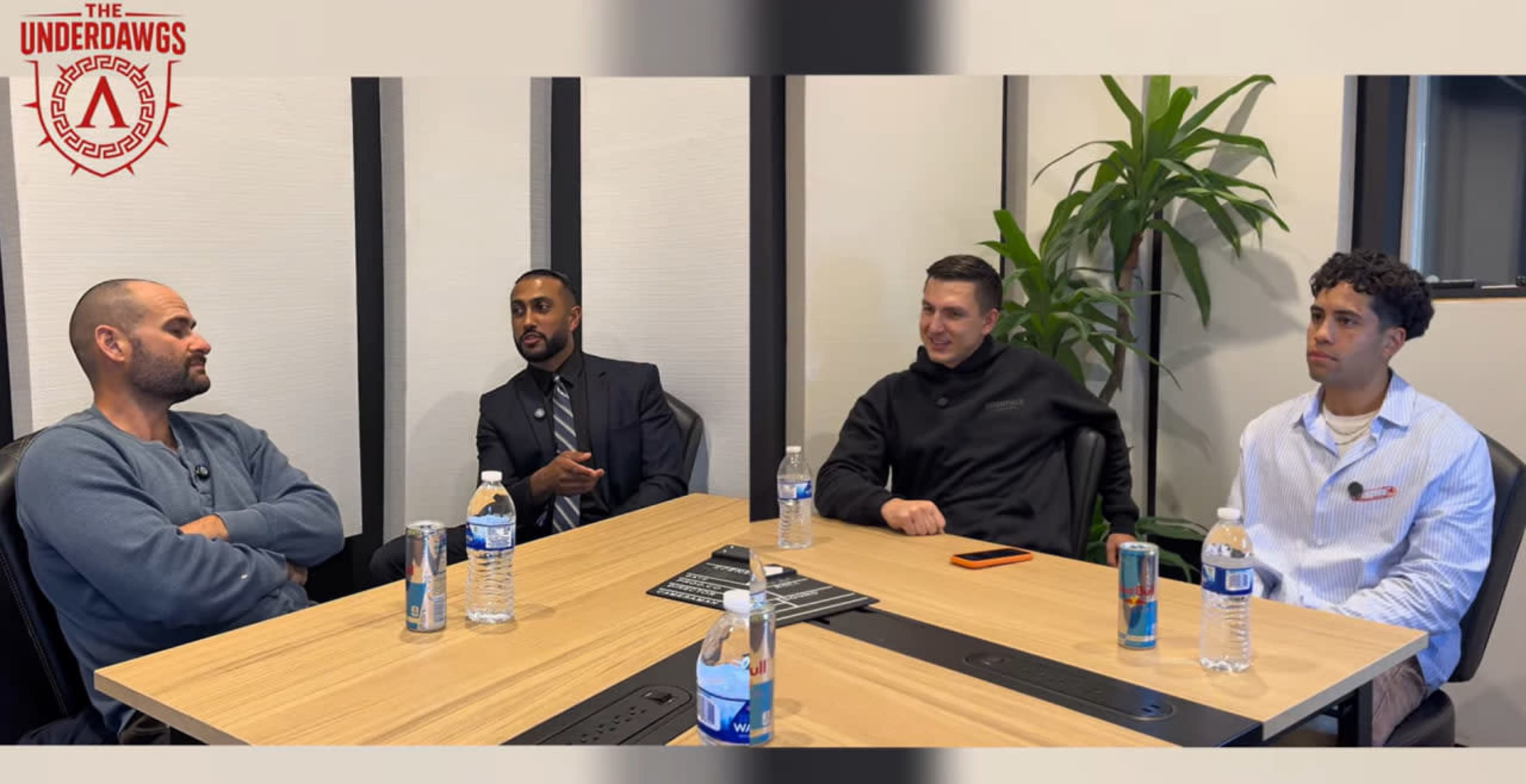 Four individuals are seated around a table in a meeting room, engaged in conversation, with water bottles placed on the table.