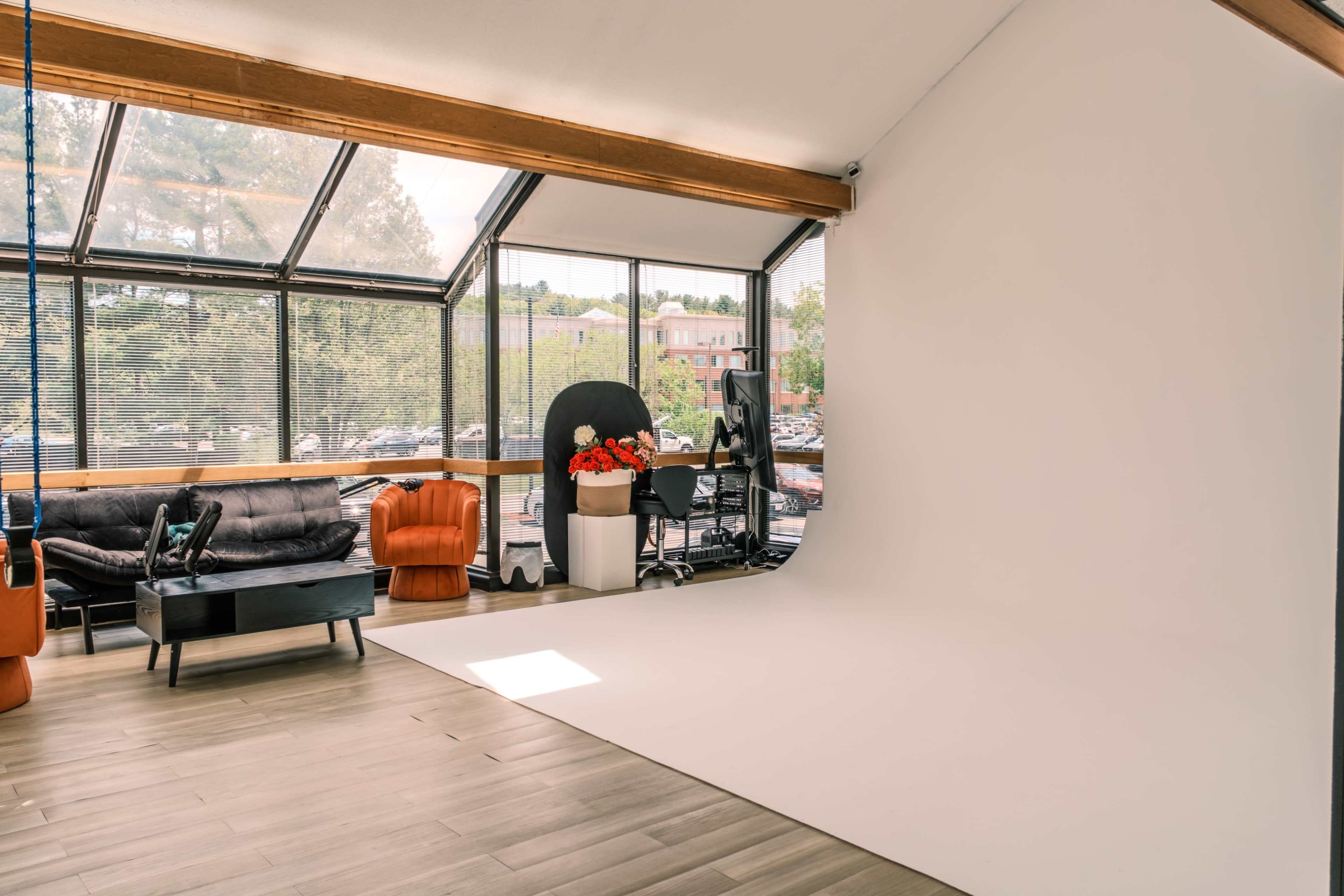 A bright studio space features a large white backdrop, a black desk chair, and modern furniture, with large windows allowing natural light.