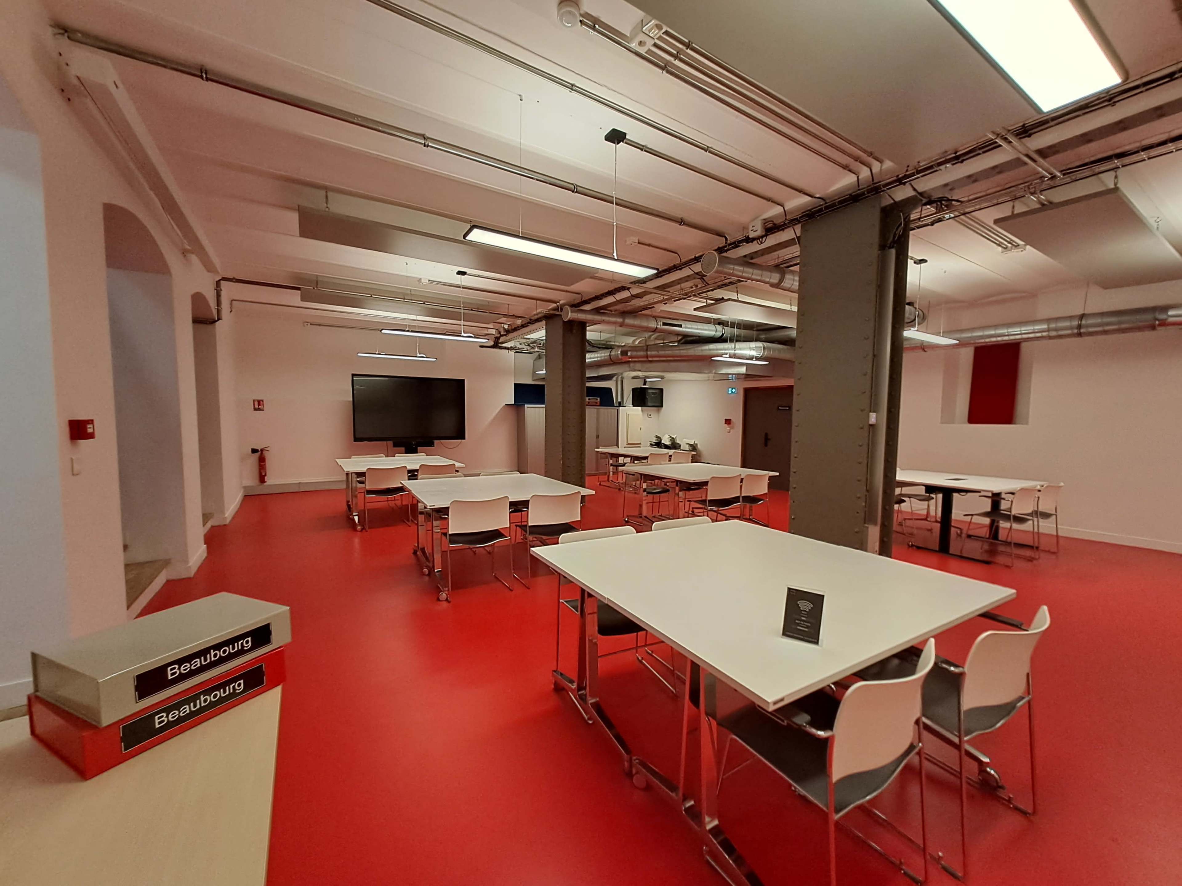A spacious room with red flooring features multiple white tables and chairs arranged throughout, with a large screen at one end.