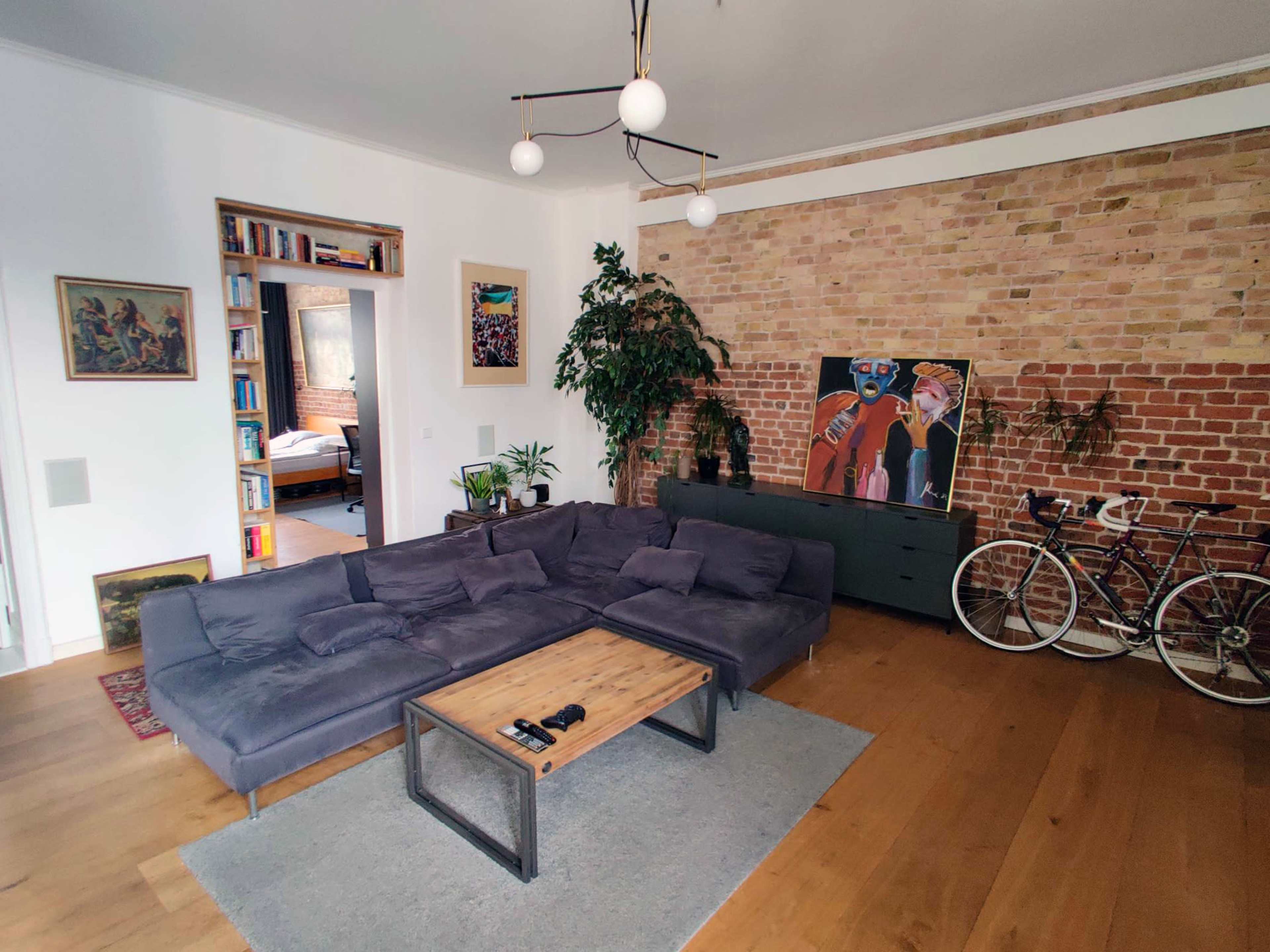 A modern living room features a gray sectional sofa, a wooden coffee table, plants, a brick wall, and a bicycle in the corner.