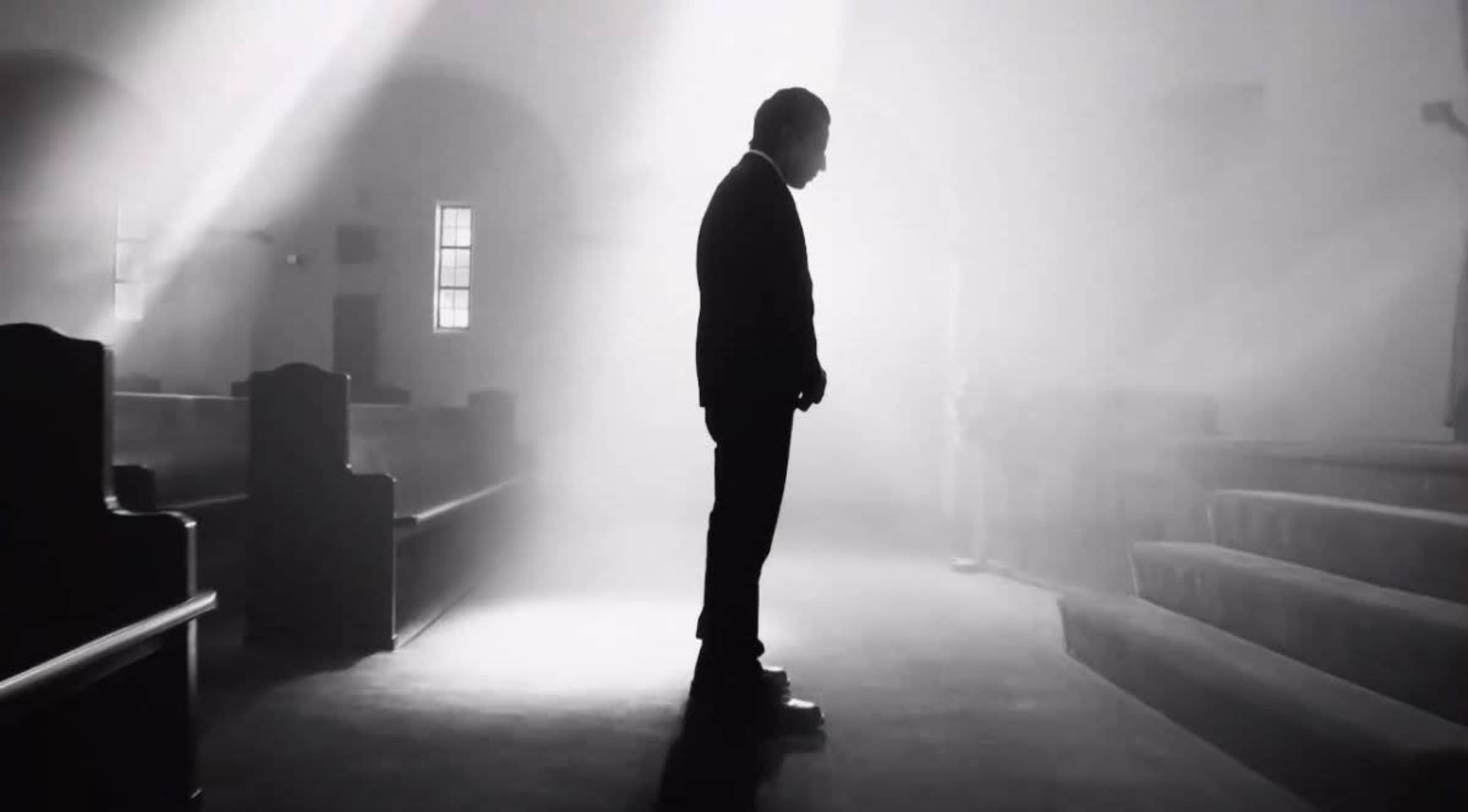 A man in a suit stands alone in a dimly lit church, surrounded by empty pews and beams of light filtering through the windows.