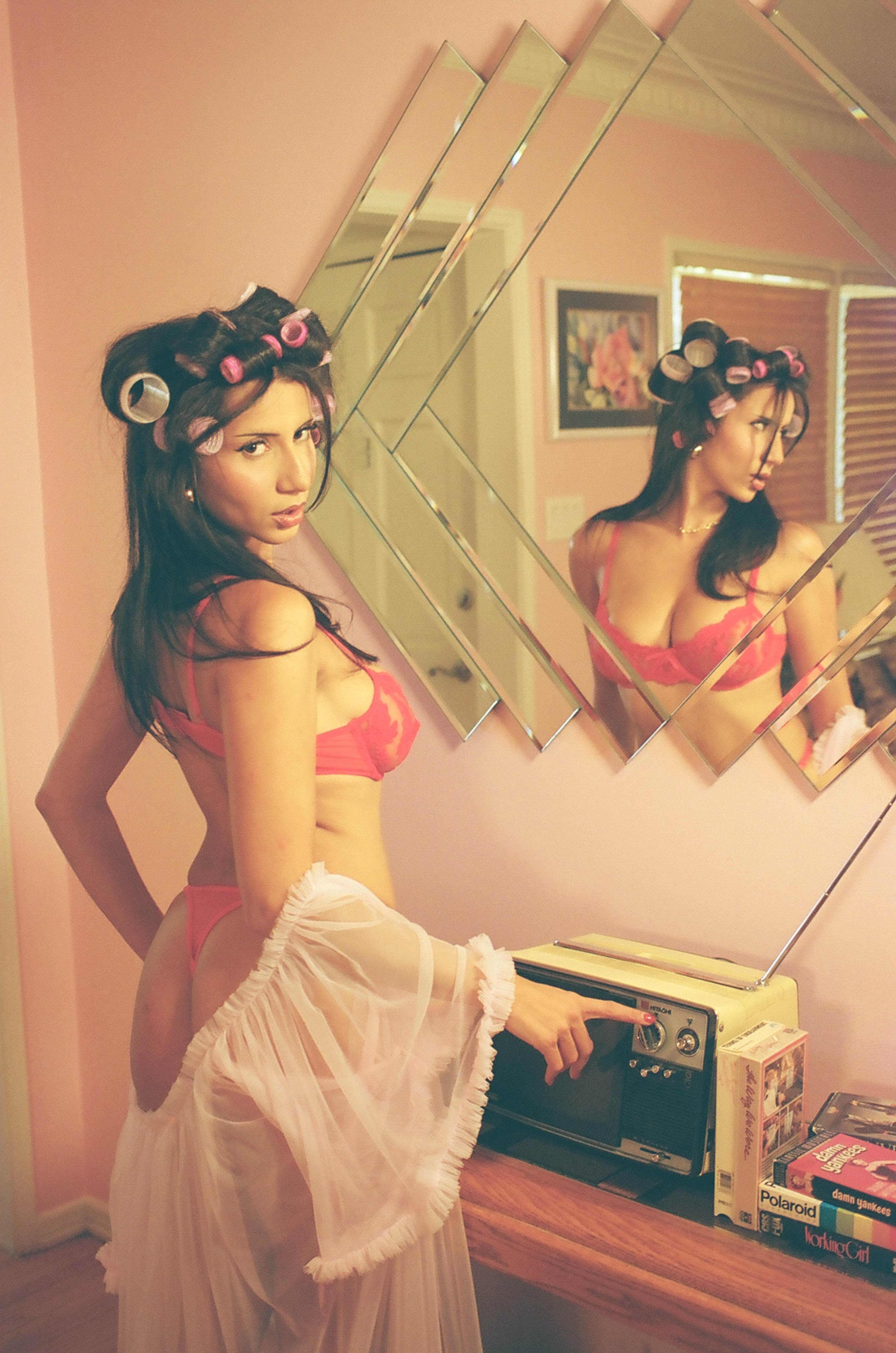 A woman wearing lingerie and hair rollers poses in front of a mirrored wall.
