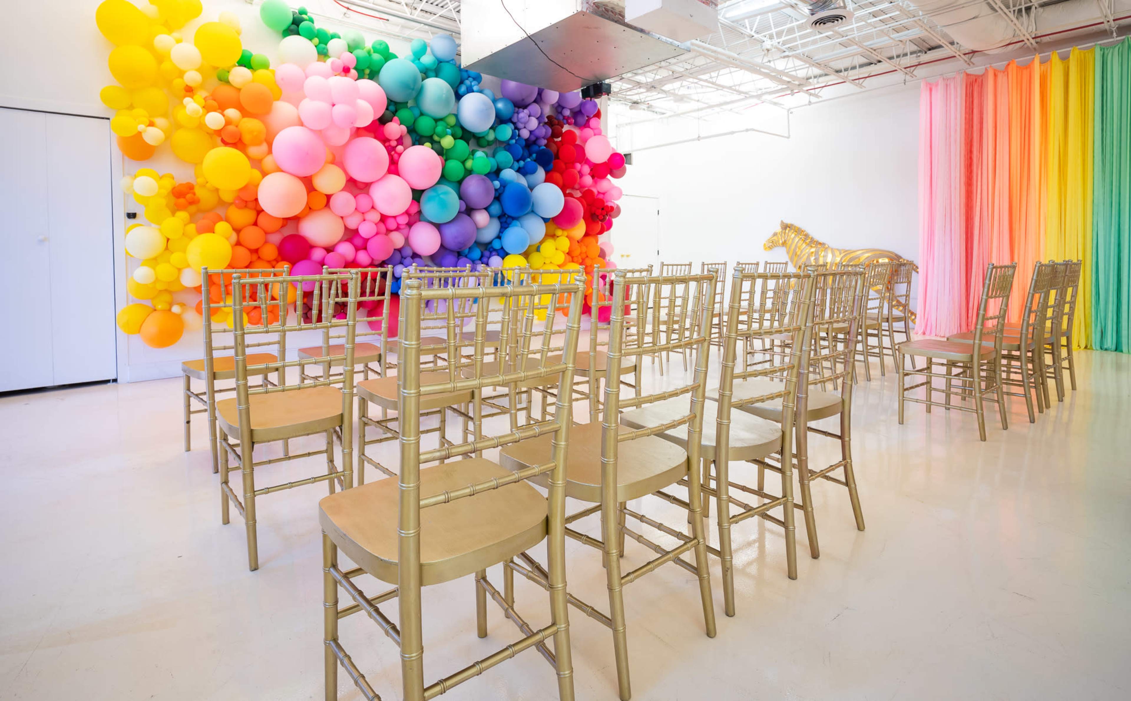 The image shows a bright room with a colorful balloon wall, golden chairs arranged in rows, and a large golden horse sculpture in the corner.