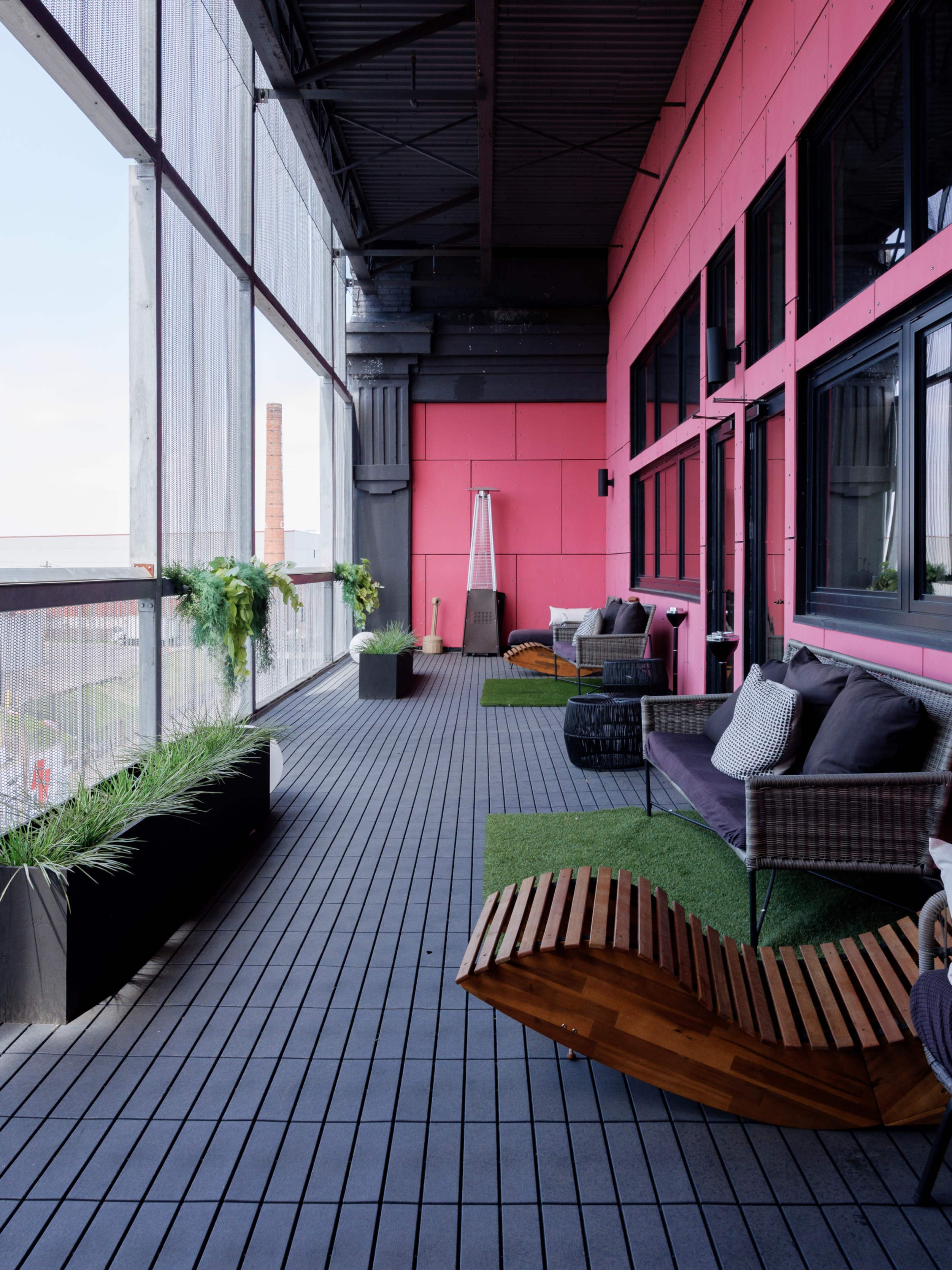 The image shows a modern balcony featuring wooden seating, potted plants, and a view of the skyline through large windows.