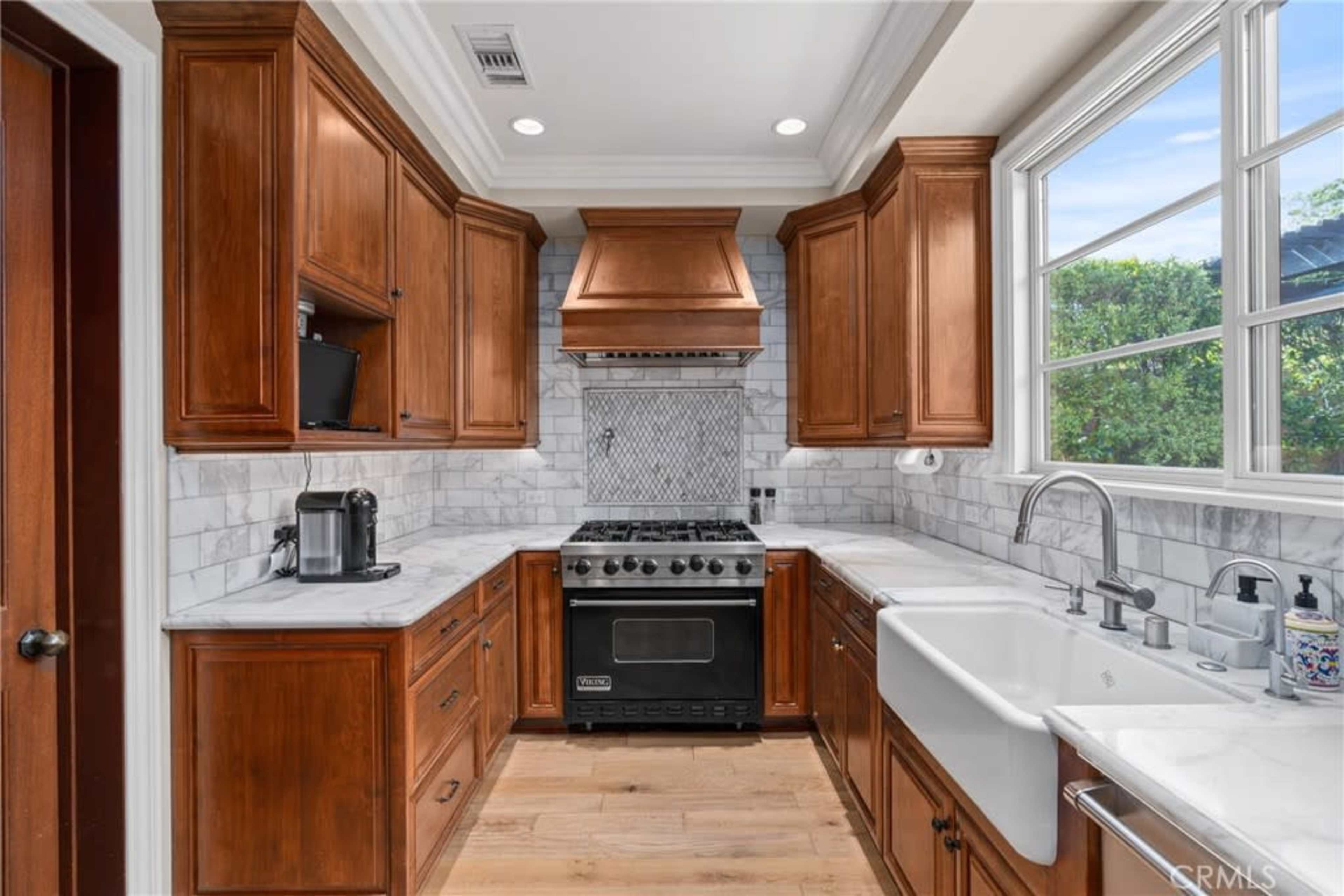 The image shows a kitchen with wooden cabinets, a stainless steel oven, a large sink, and a window allowing natural light to enter.