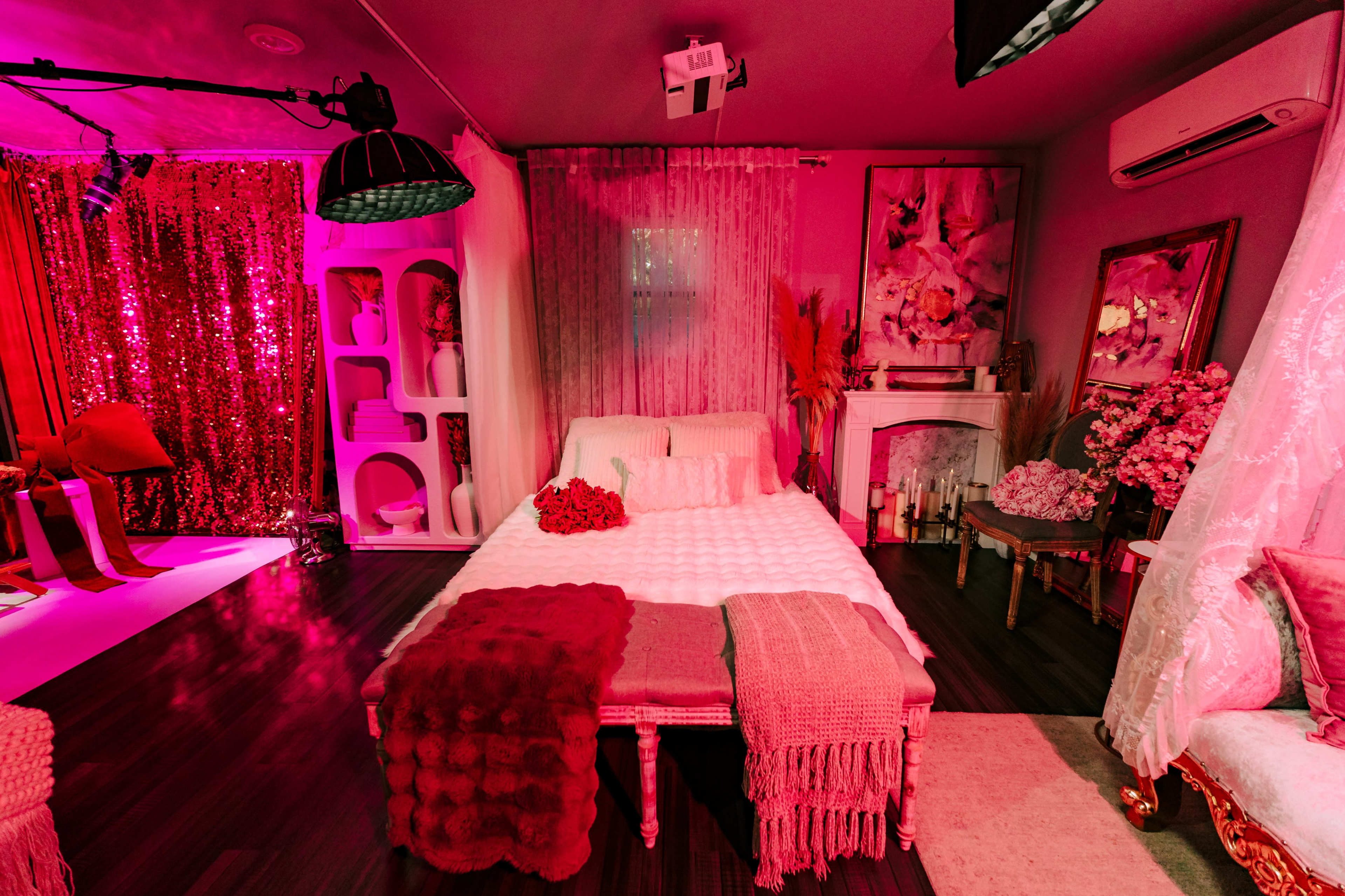The image depicts a decorated bedroom with a bed covered in white linens, surrounded by pink lighting, floral artwork, and soft furnishings.