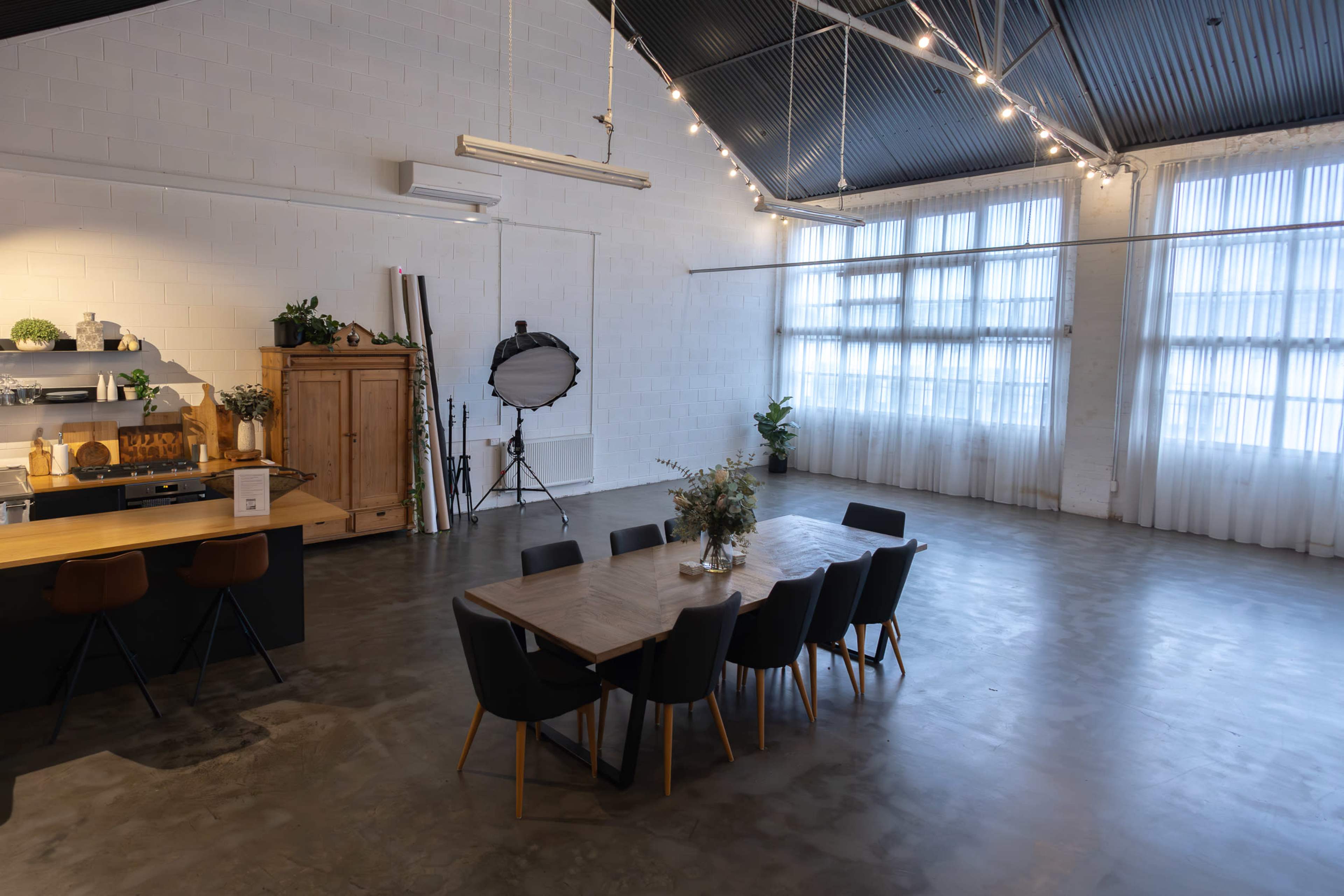 The image shows a spacious, well-lit interior with a dining table surrounded by chairs, a kitchen area, and large windows adorned with sheer curtains.