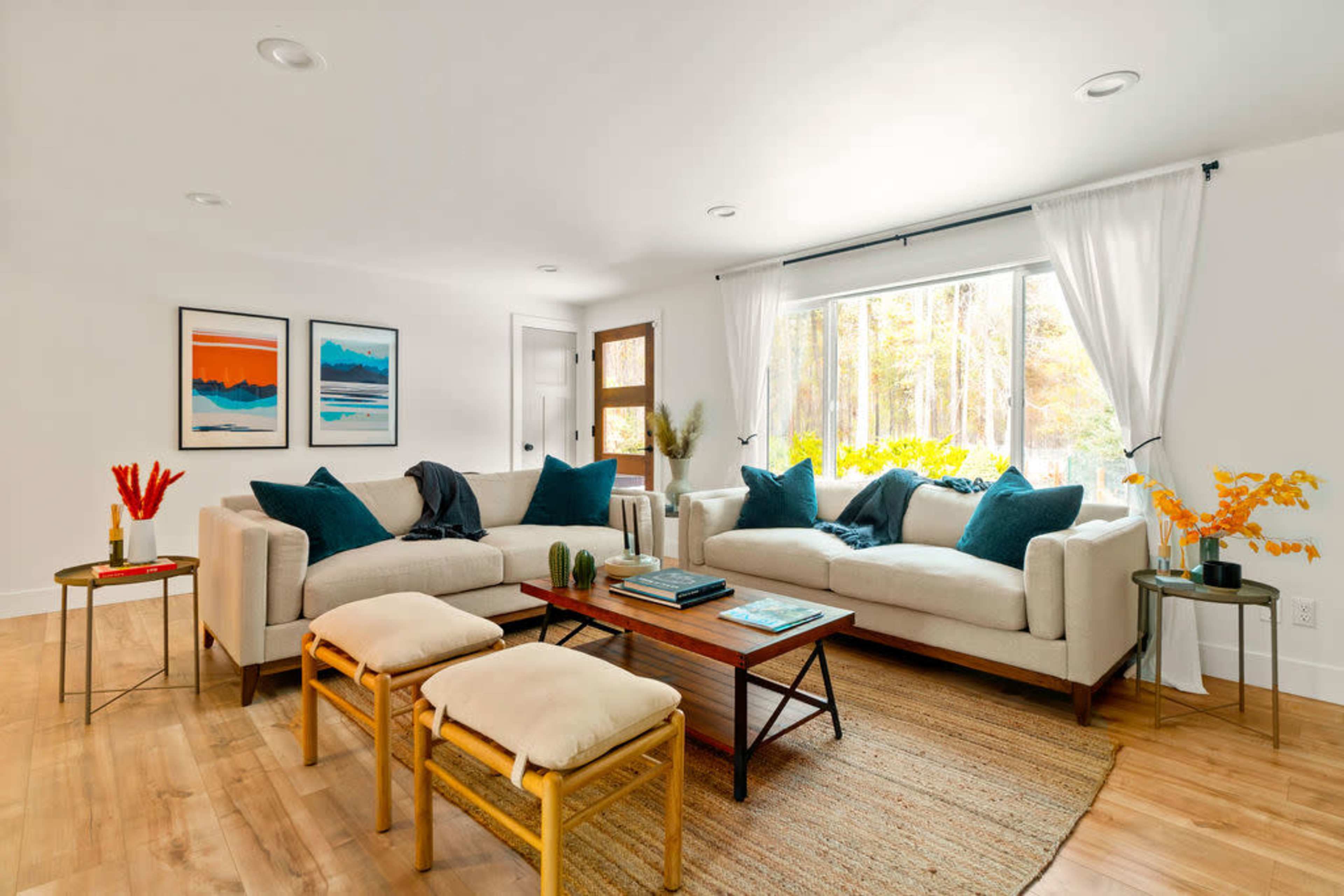 A bright living room features a light-colored sofa with teal cushions, a coffee table with books and plants, and large windows letting in natural light.