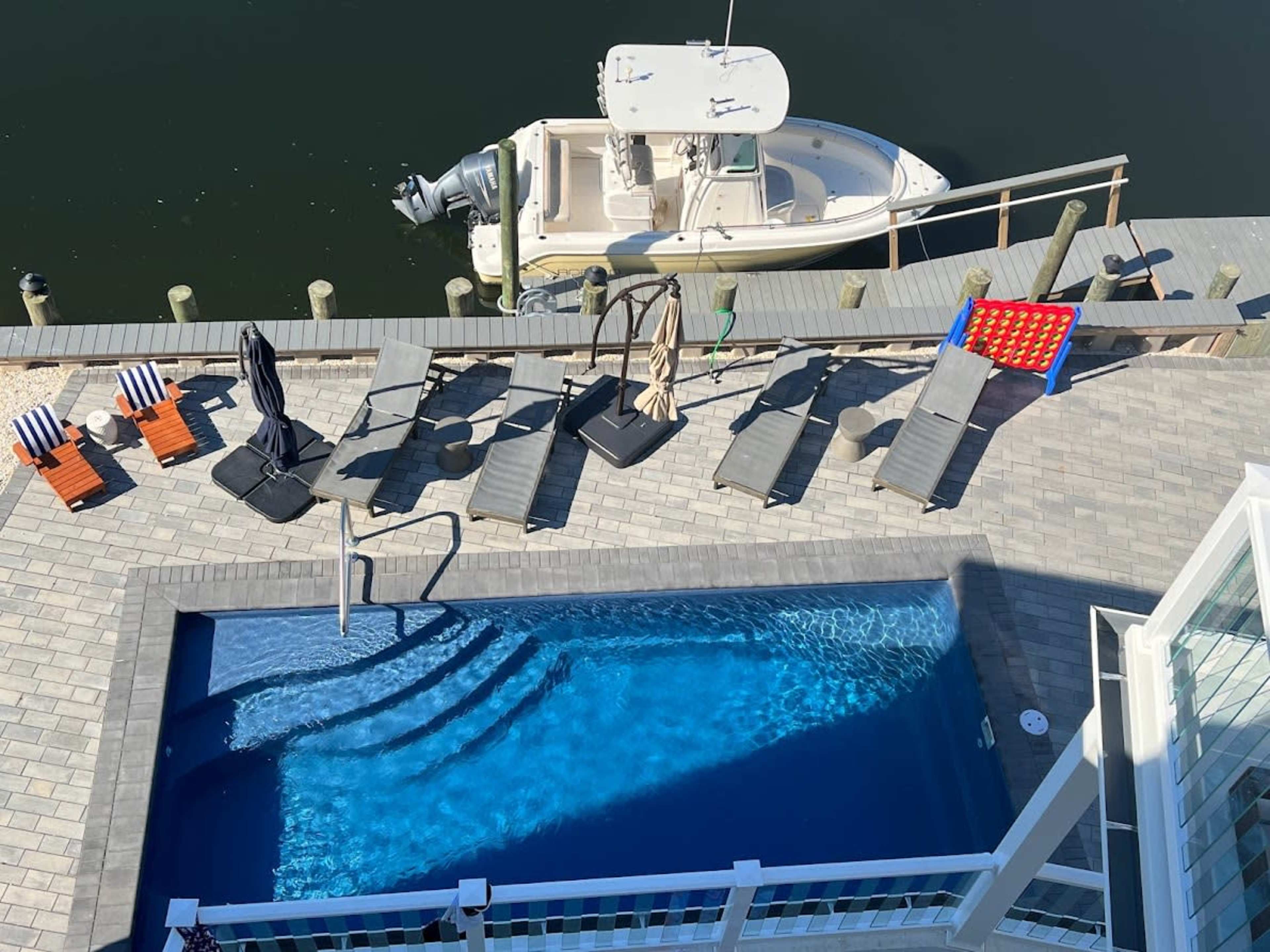 The image shows a poolside area with several lounge chairs arranged around a small swimming pool and a boat docked nearby on the water.