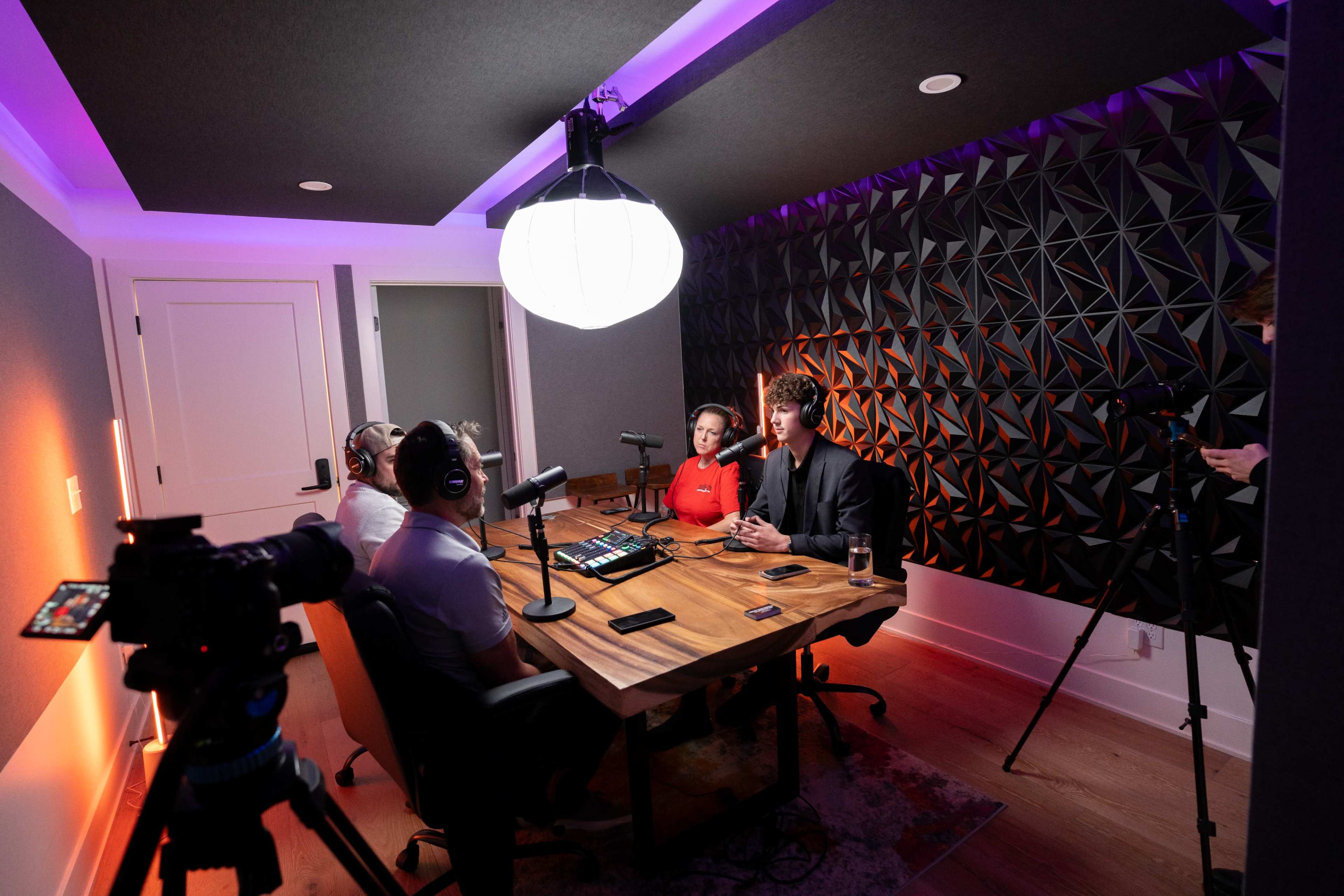 A group of four people sits around a wooden table in a podcast recording studio, equipped with microphones and recording equipment, while a fifth person stands nearby with a camera.