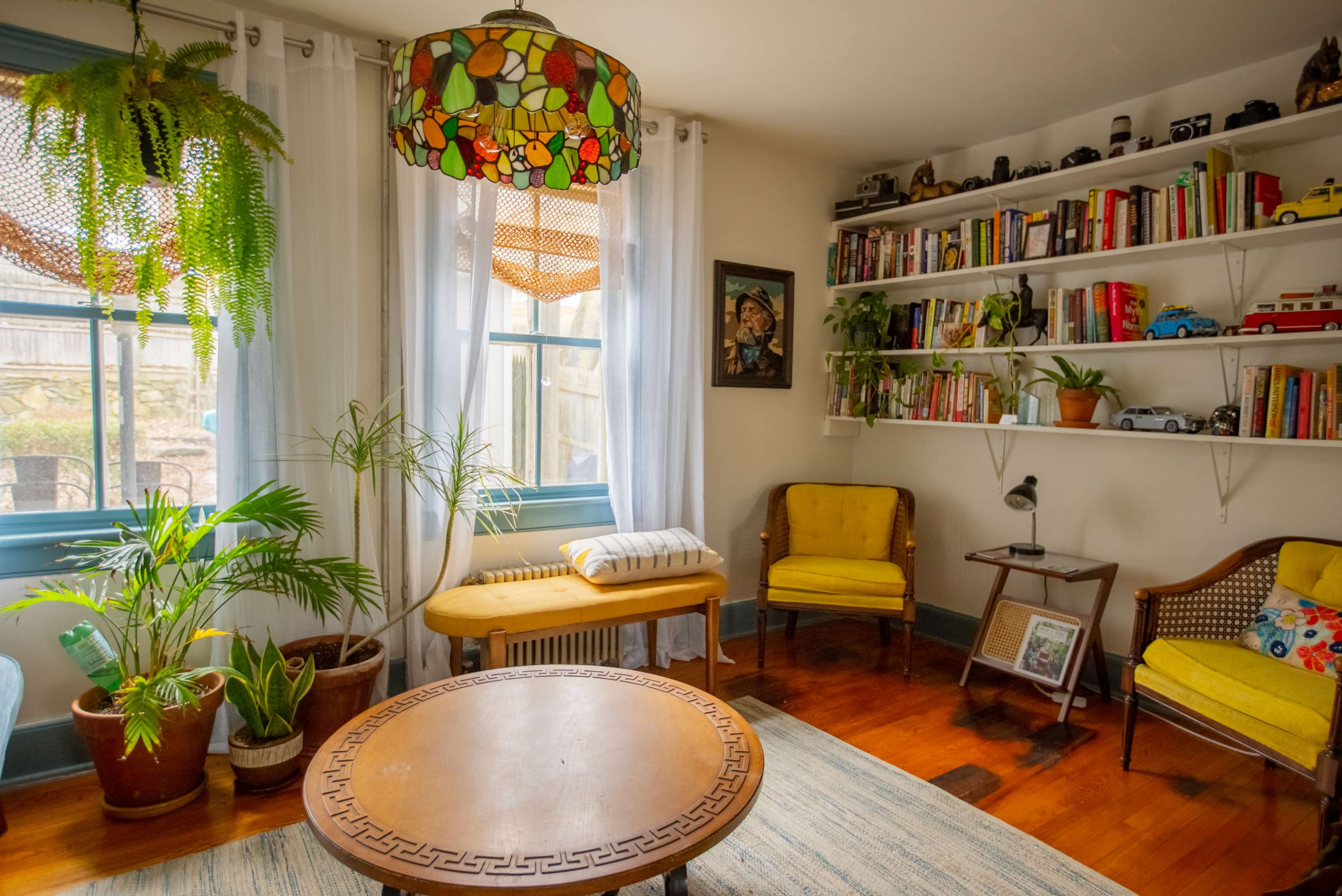 The image shows a cozy living room with a round wooden table, yellow chairs, a shelf filled with books and collectibles, and large windows featuring light curtains and indoor plants.