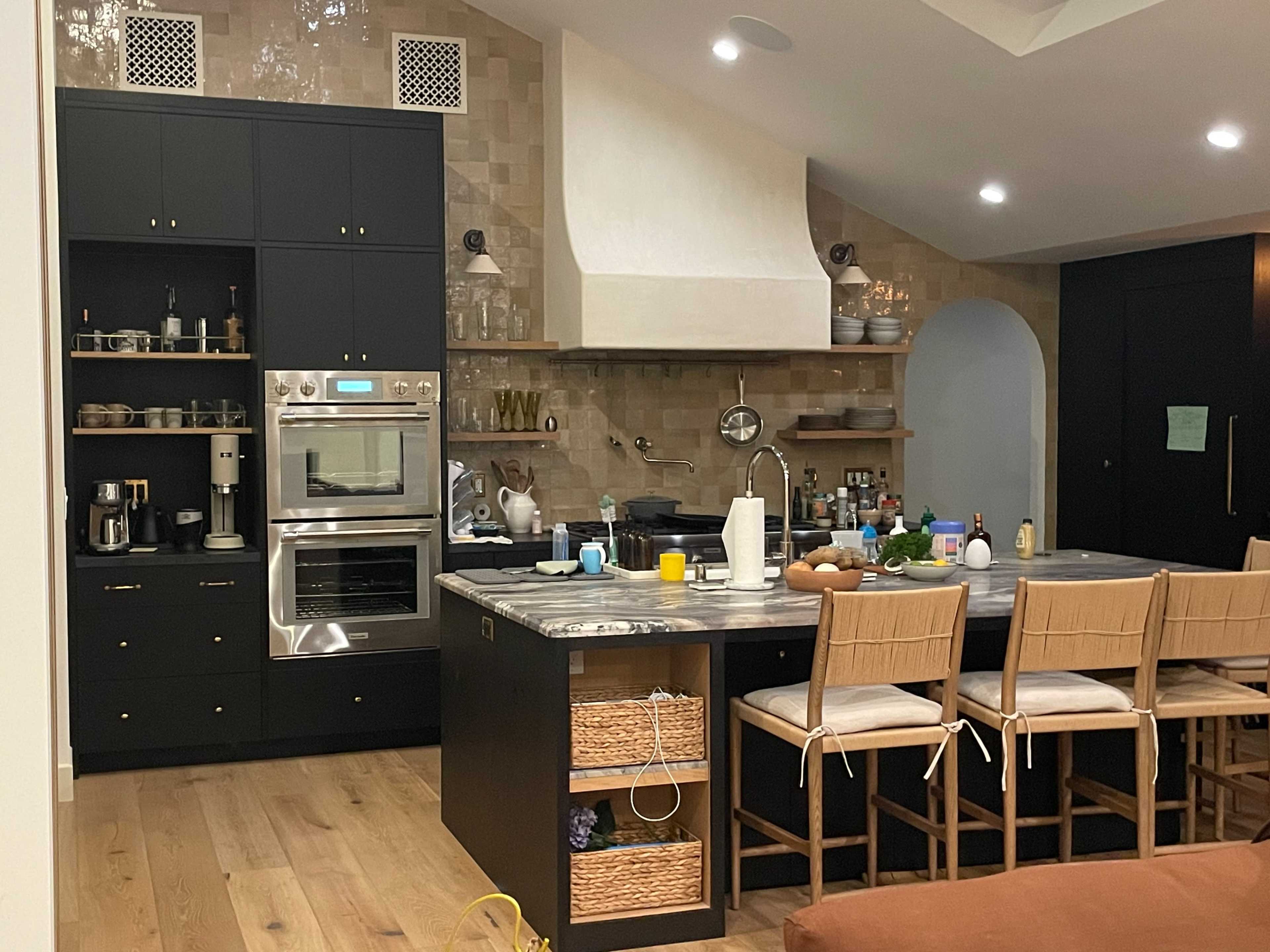 The image shows a modern kitchen featuring a large island with seating, built-in ovens, and various kitchen appliances organized on dark cabinetry and a light-colored backsplash.