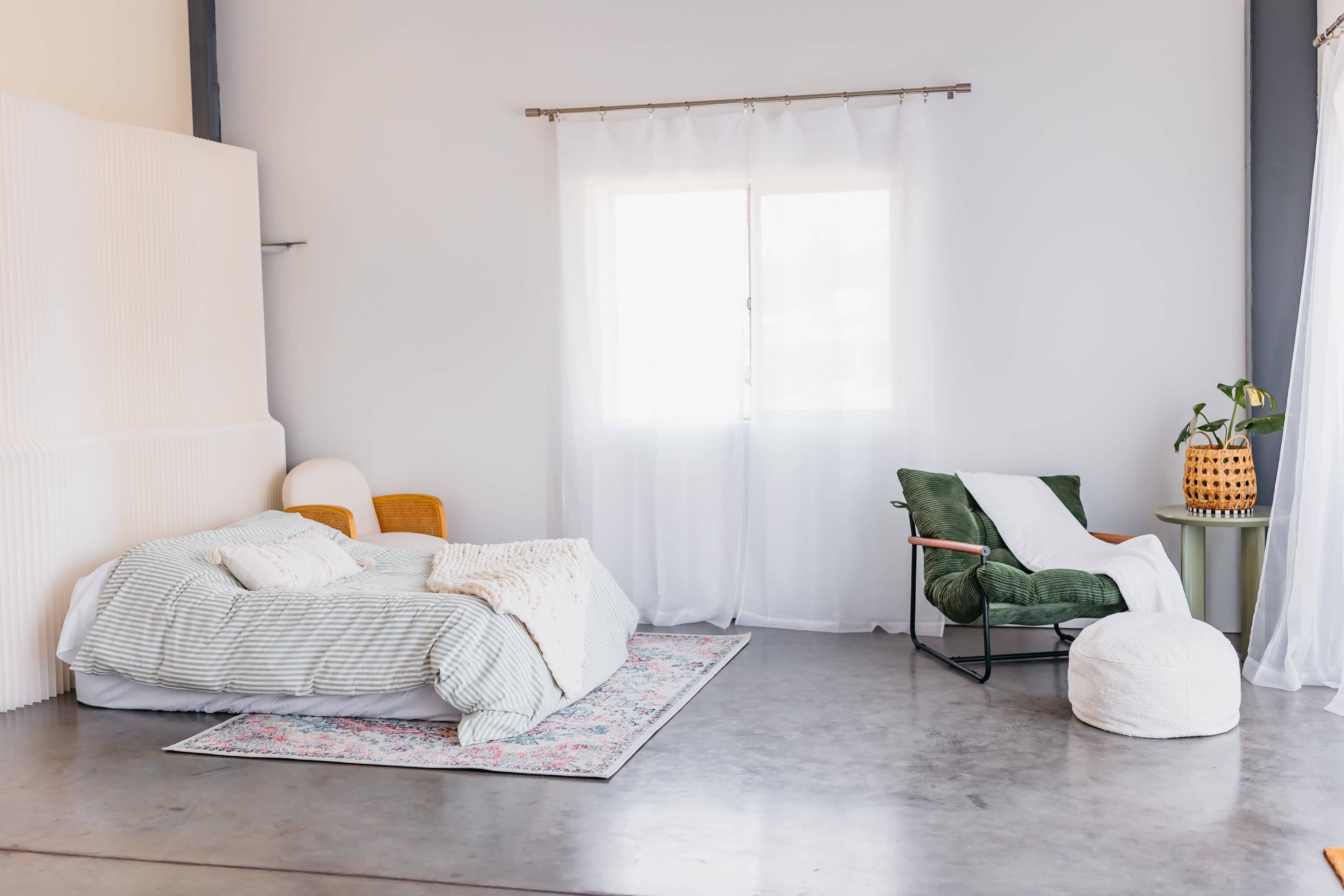 The image shows a minimalist room featuring a bed with layered bedding, a green chair, a round pouf, and large windows covered with sheer white curtains.