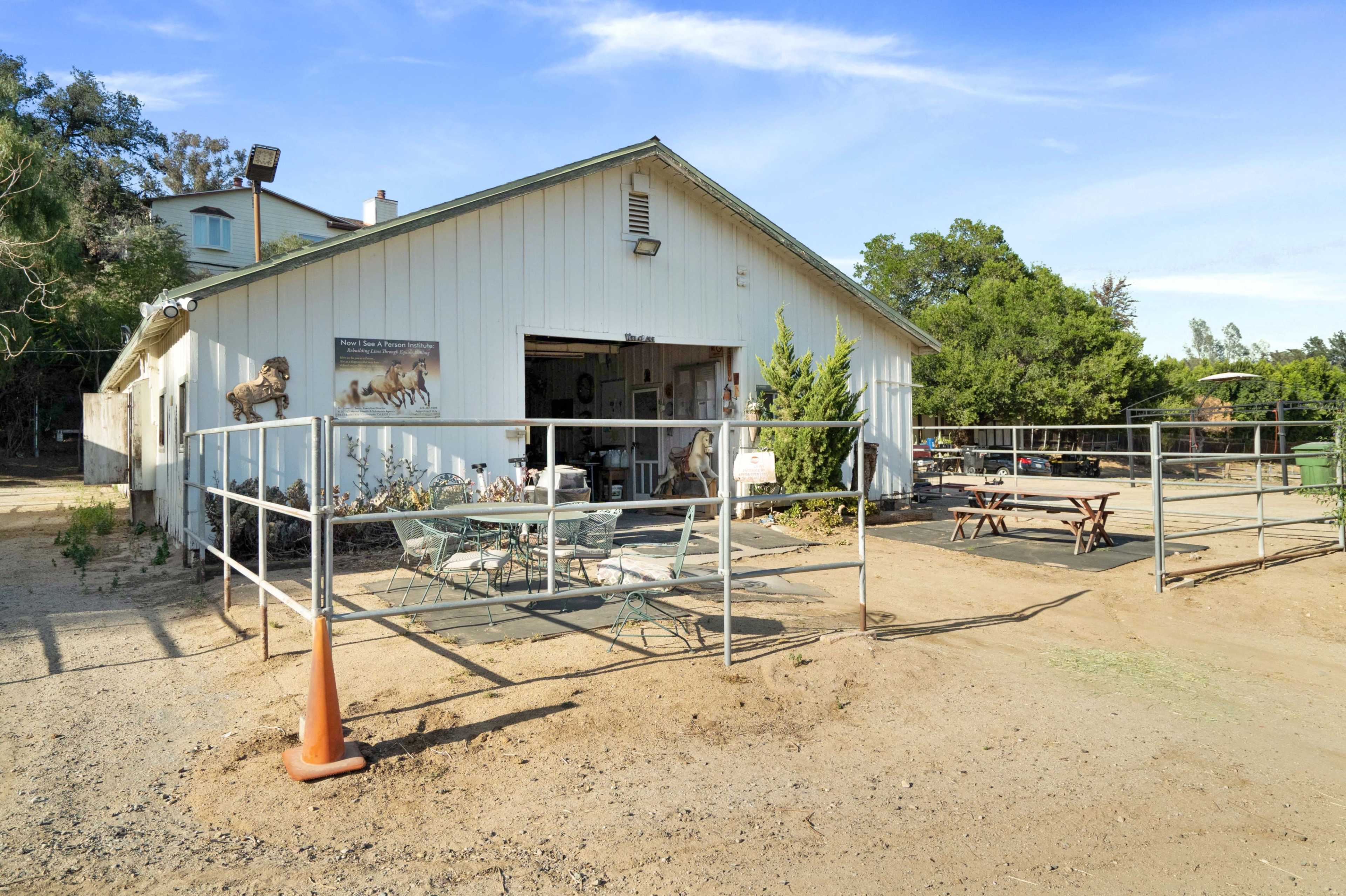 The image shows a white barn-style building with outdoor seating and a fenced area in a rural setting.