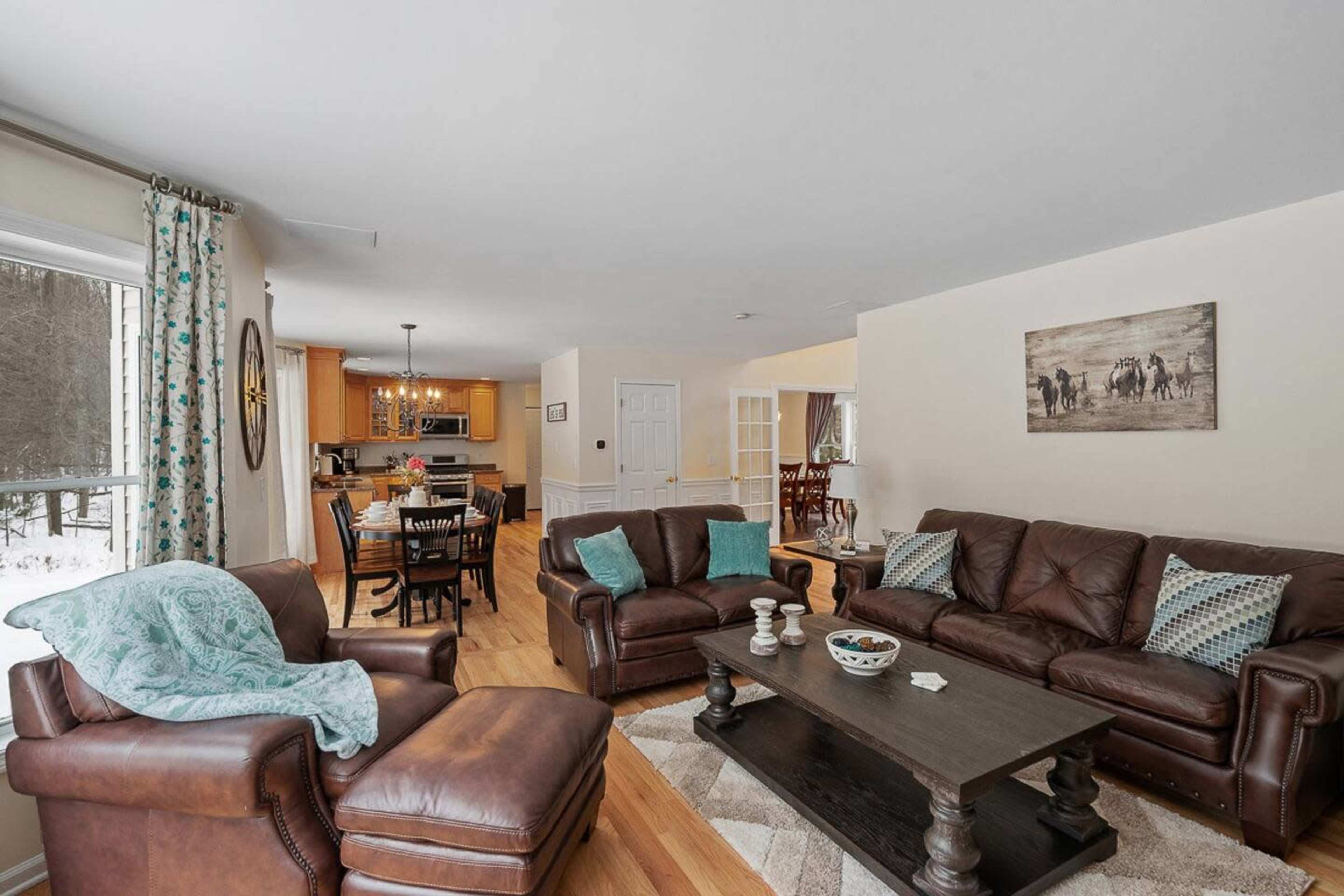 The living room features brown leather sofas arranged around a dark coffee table, with a view of a dining area and kitchen in the background.