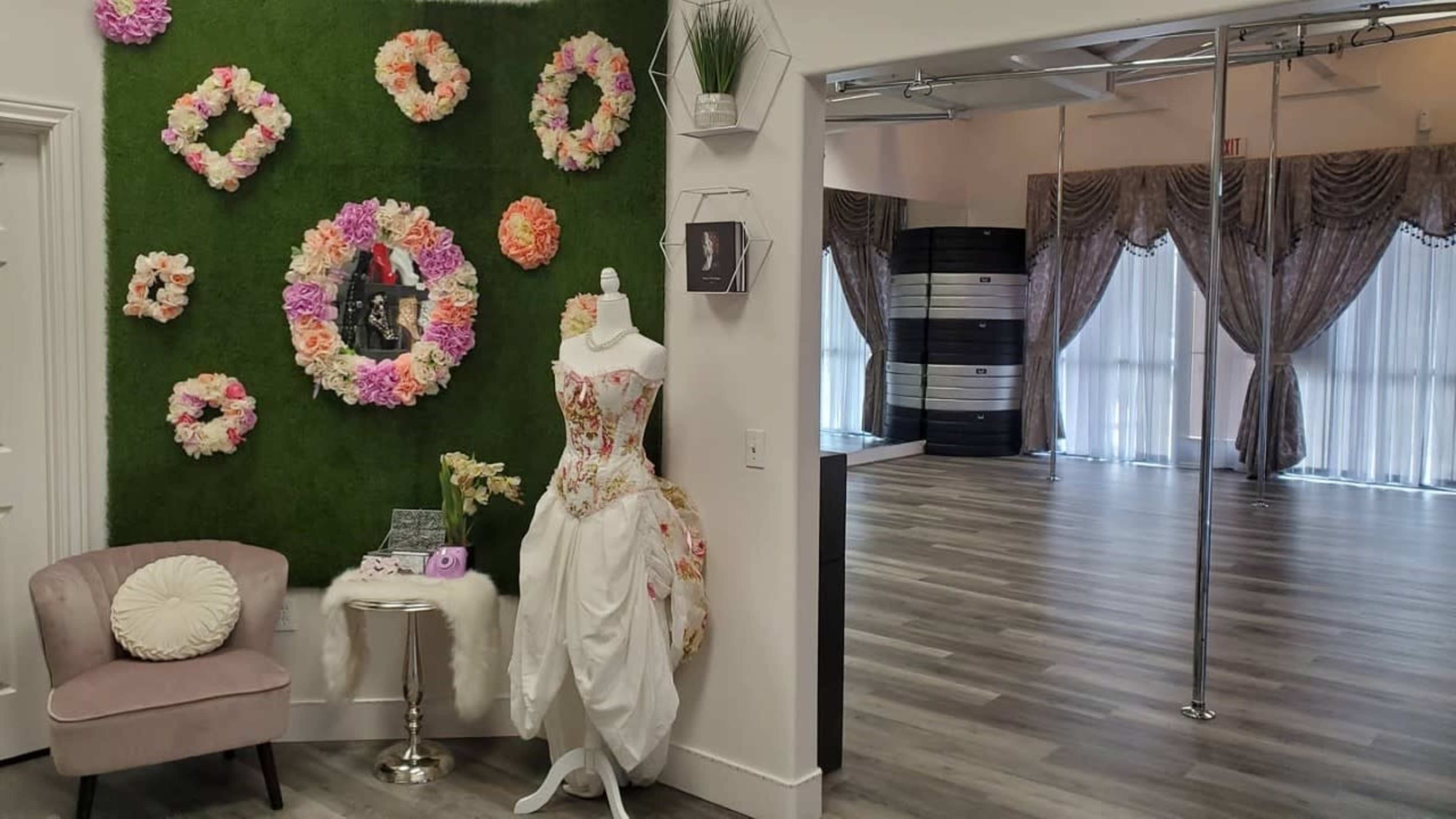 The image shows a bridal display featuring a white dress on a mannequin beside a floral wall, with a spacious room in the background designed for events.