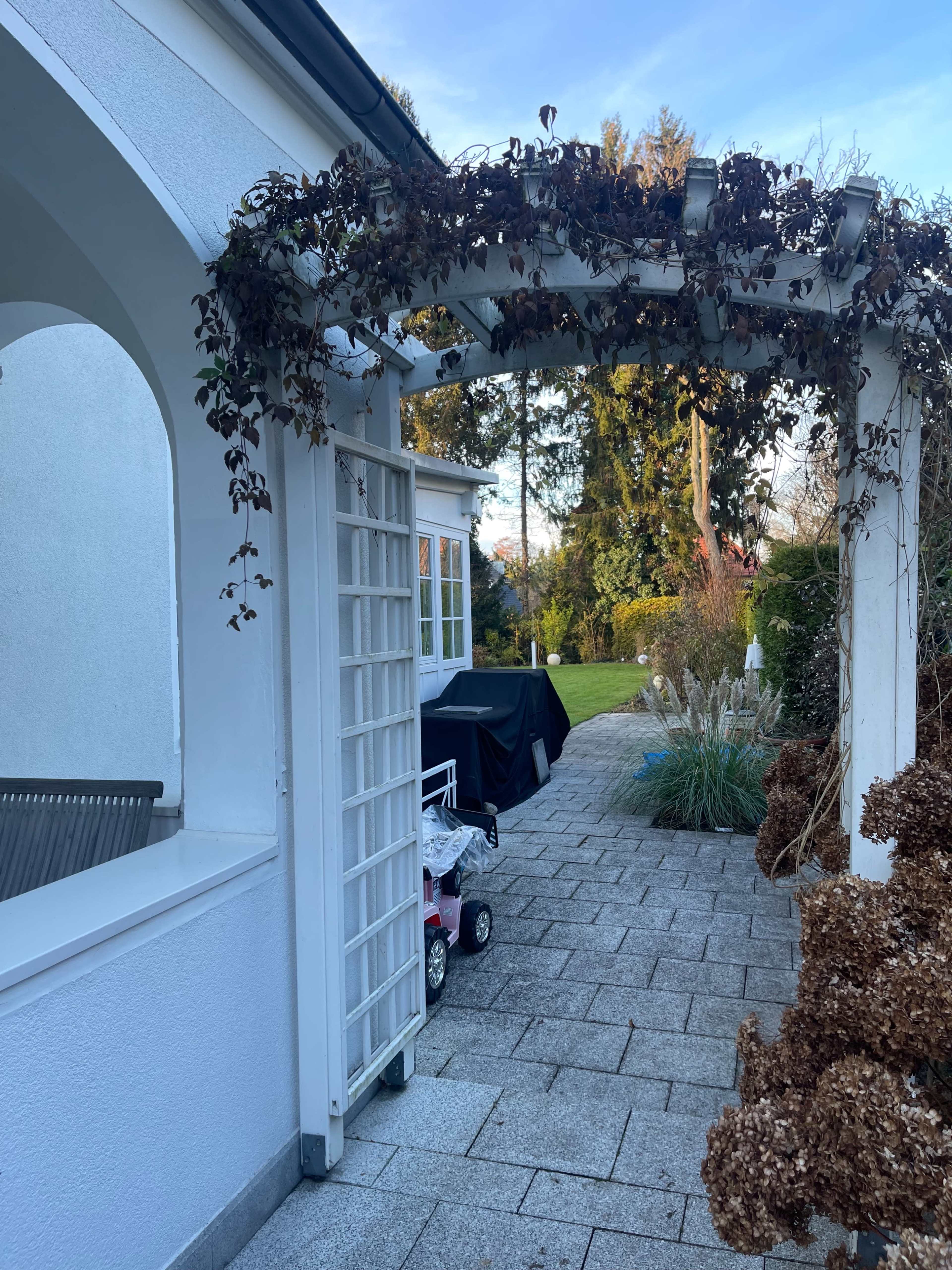 A white trellis arch leads to a paved patio area with a barbecue grill and a garden in the background.