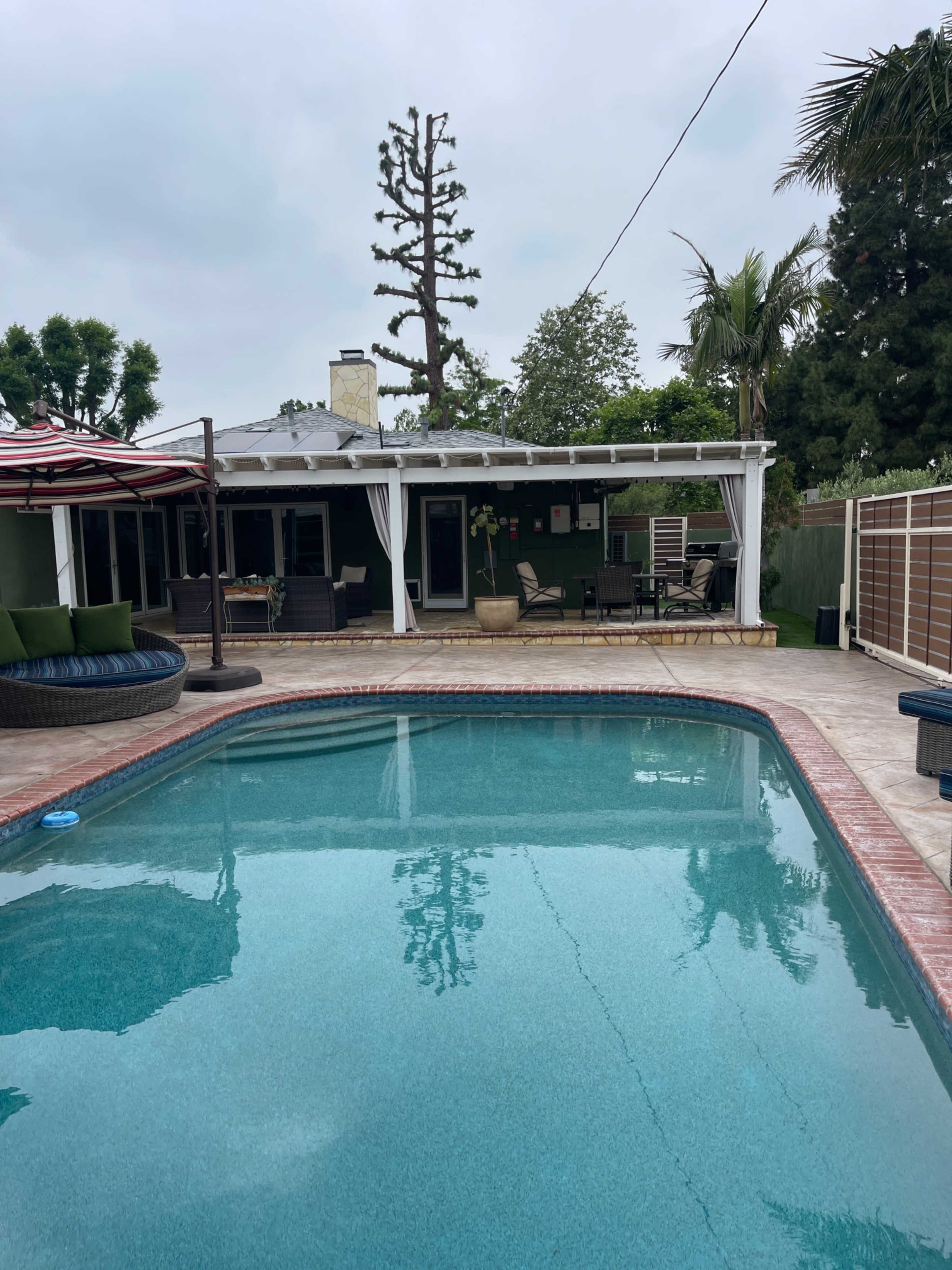 A clear swimming pool reflects the surrounding patio and house, which features a covered seating area and tall trees in the background.