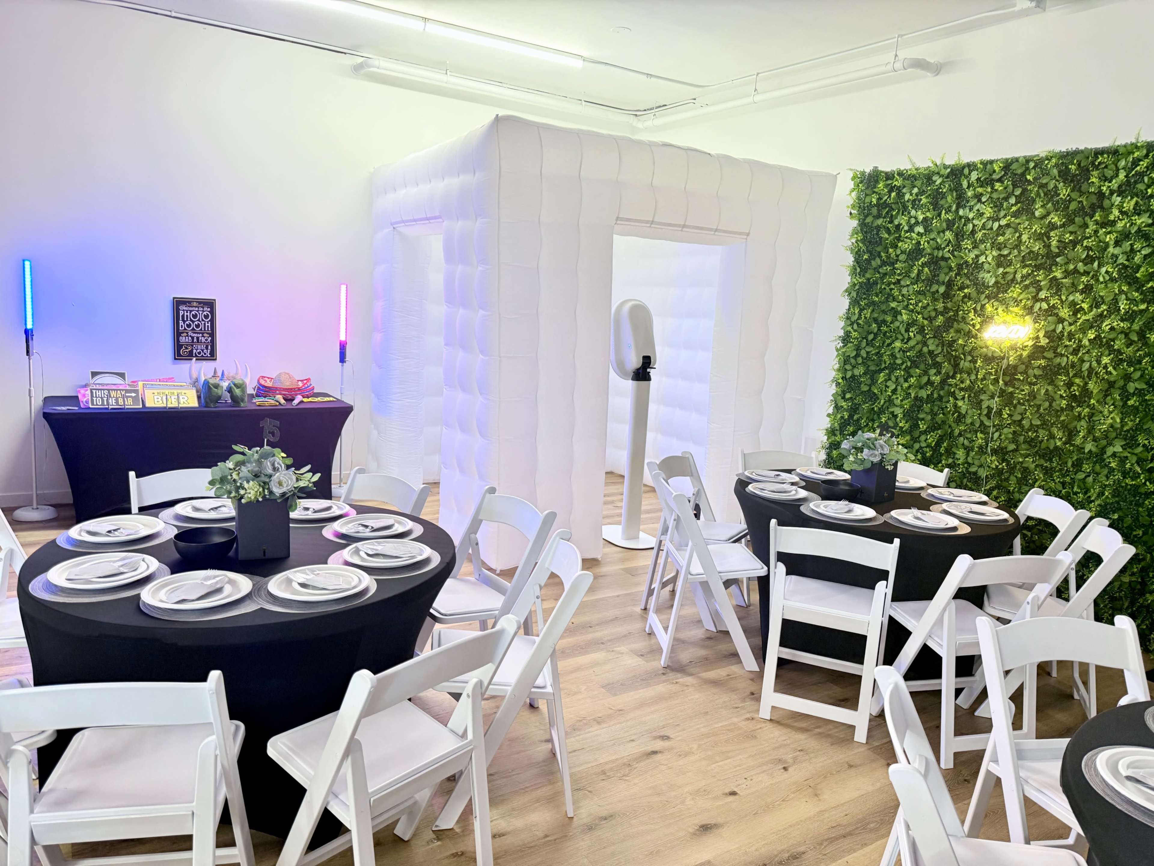 The image shows a decorated event space with black tables set for dining, white chairs, and a backdrop featuring a lush green wall and a white inflatable structure.