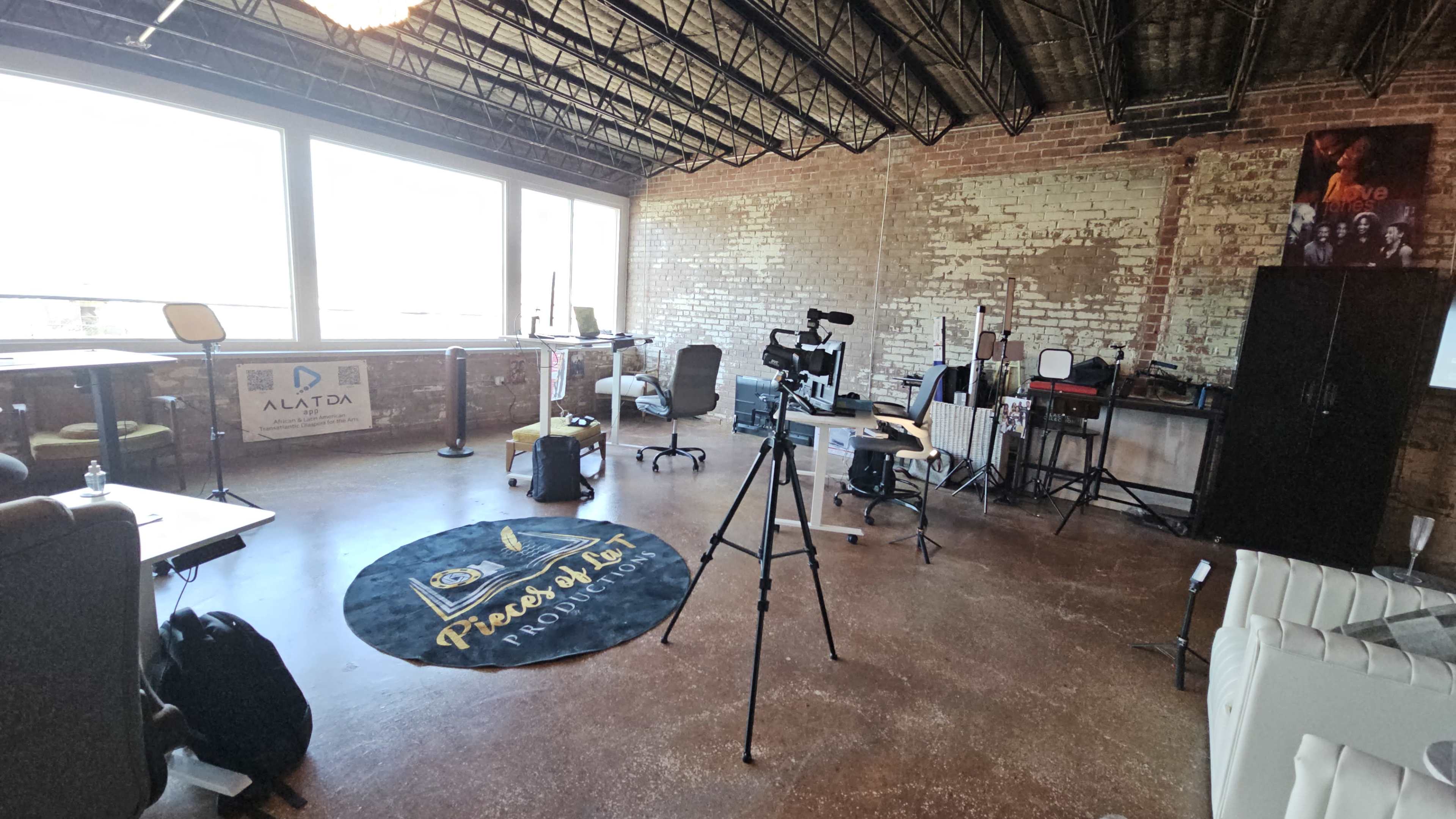 The image shows a spacious, industrial-style office setting with various workstations, a camera on a tripod, and large windows letting in natural light.