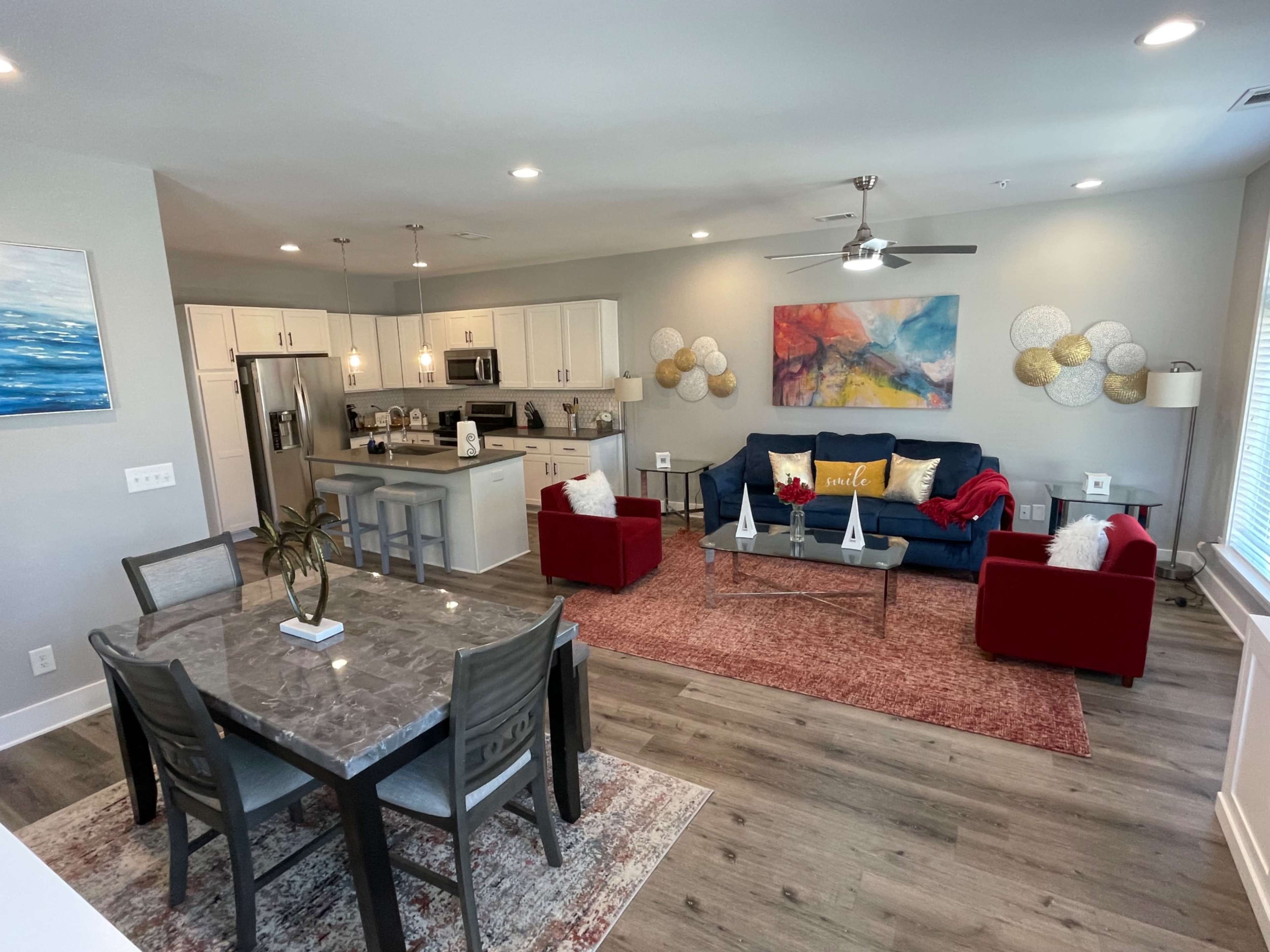 The image shows a modern open-concept living area featuring a dining table, a blue sofa, red armchairs, and a kitchen in the background.