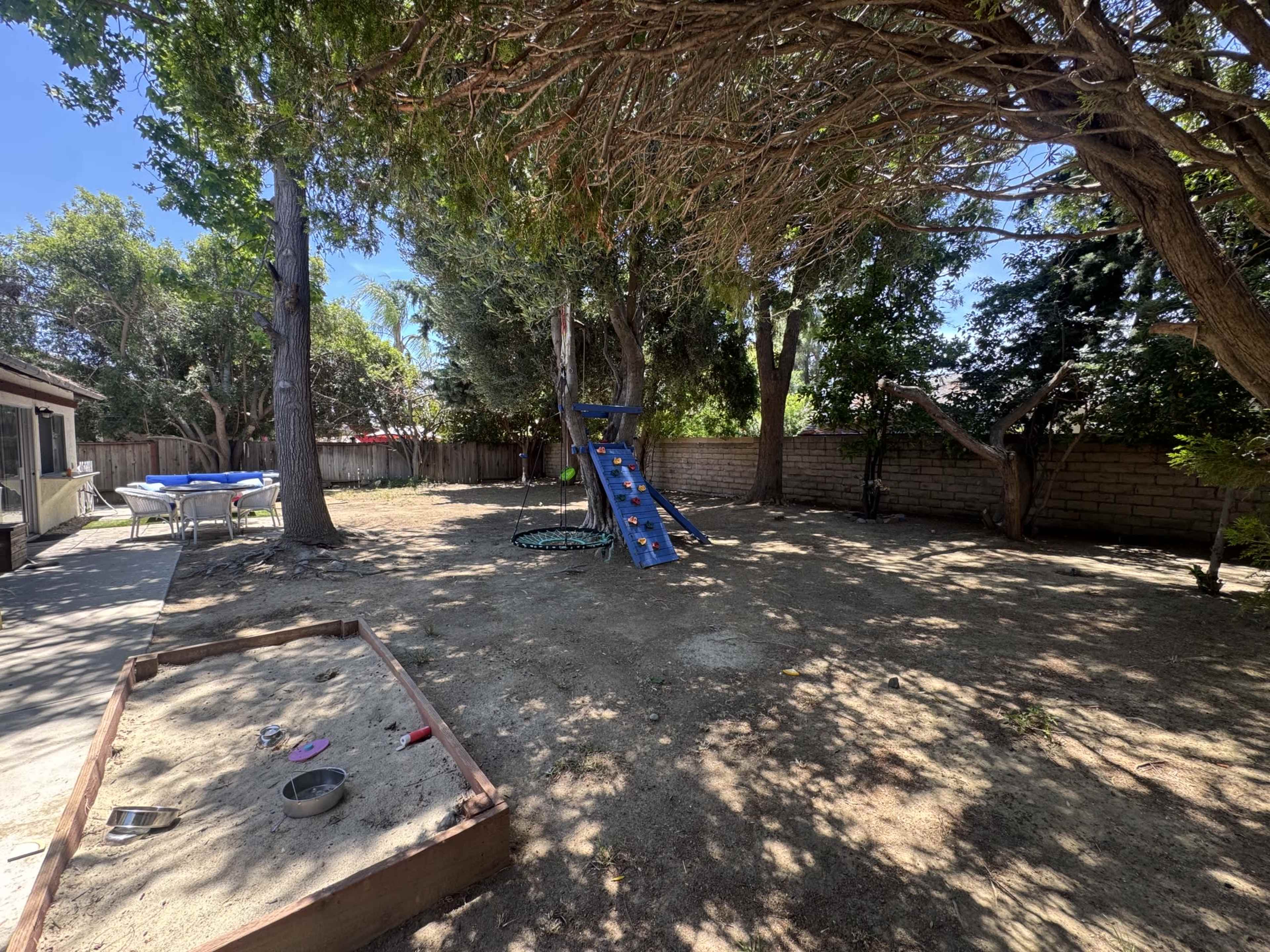 The image shows a backyard with a play structure, a sandbox, and several trees under a clear blue sky.
