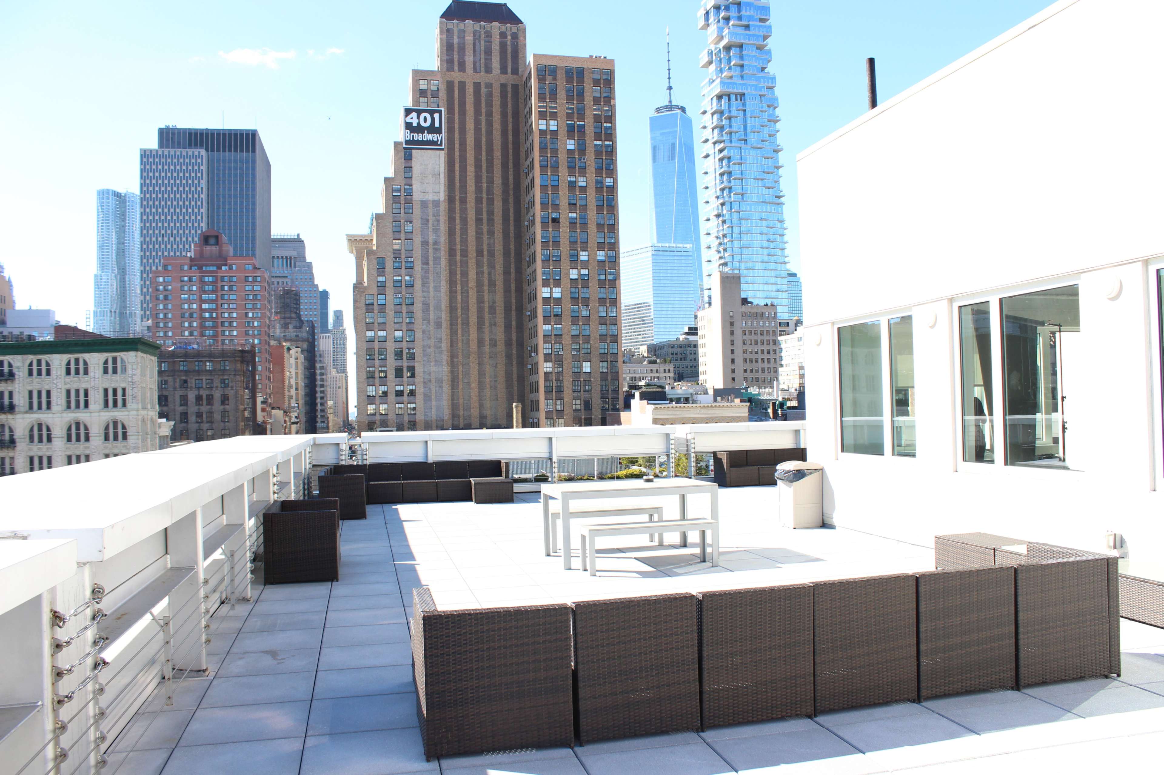 The image shows a rooftop terrace with a few outdoor furniture pieces, overlooking tall city buildings.