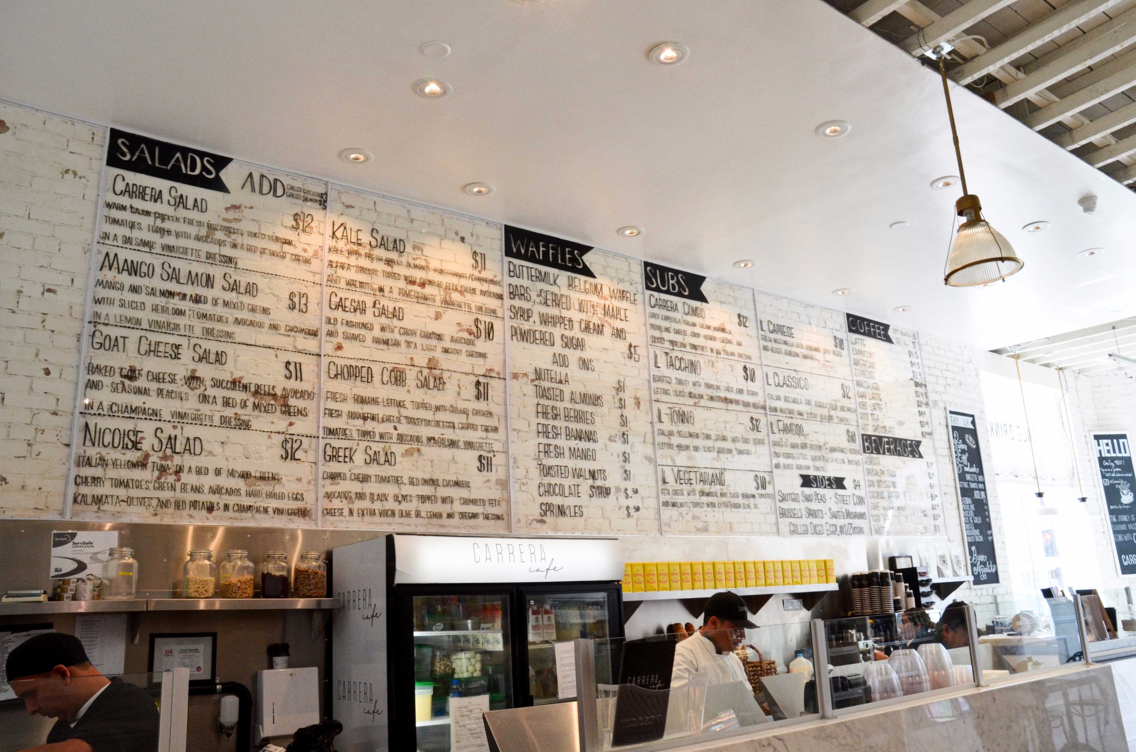 A menu board featuring various food items, including salads, waffles, and subs, displayed on a white brick wall in a restaurant.