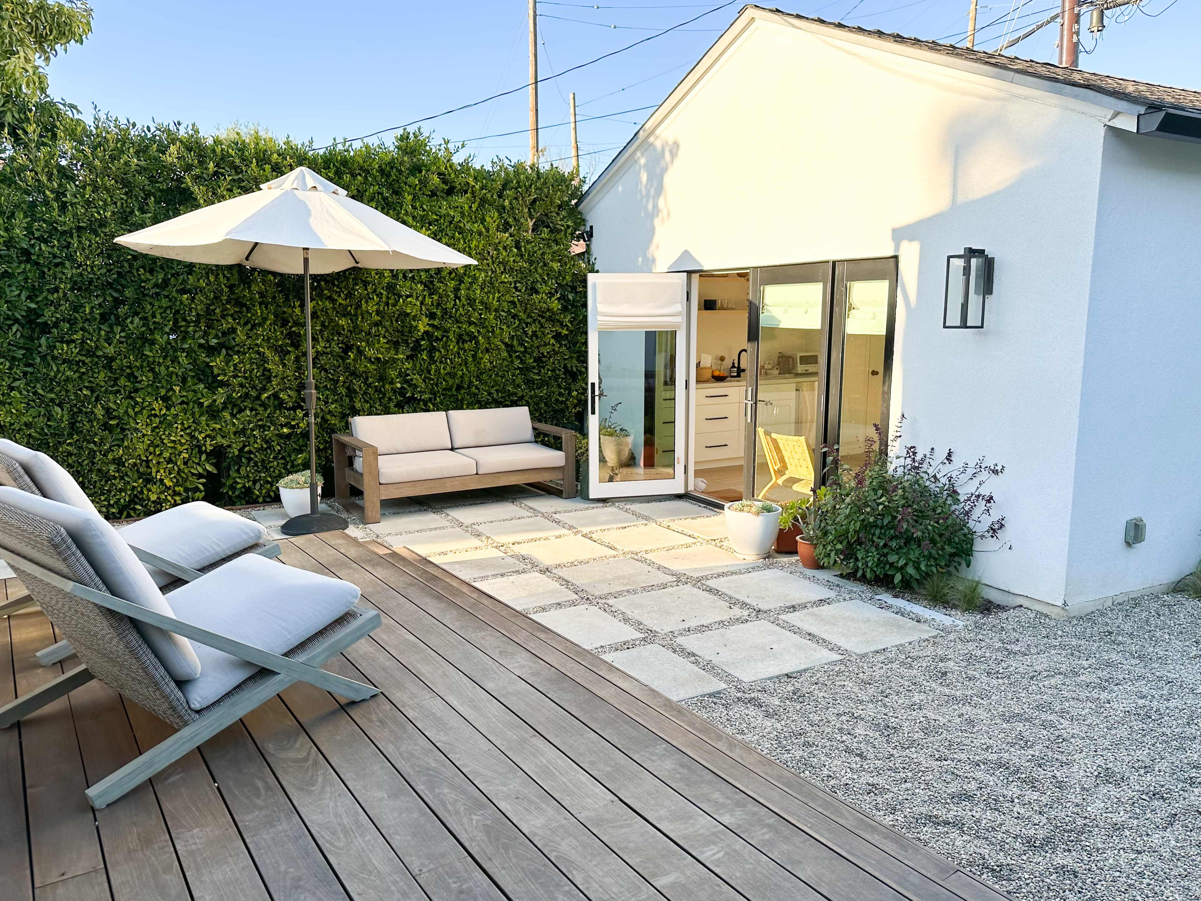 A modern outdoor space features a wooden deck with lounge chairs, a sofa, and a small patio surrounded by greenery and a white house.