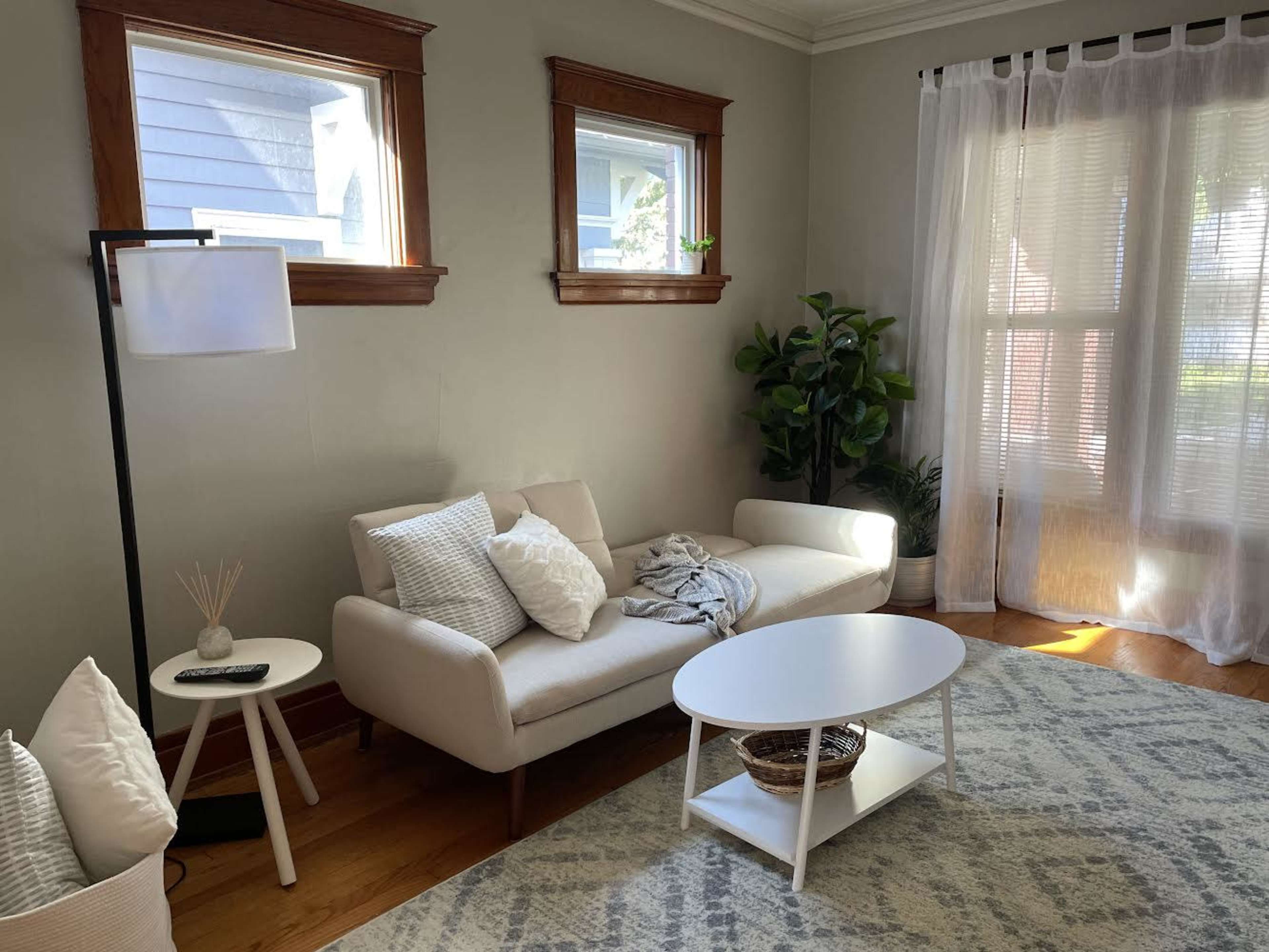 A cozy living room with a white sofa, a small round table, and large windows allowing natural light to enter.