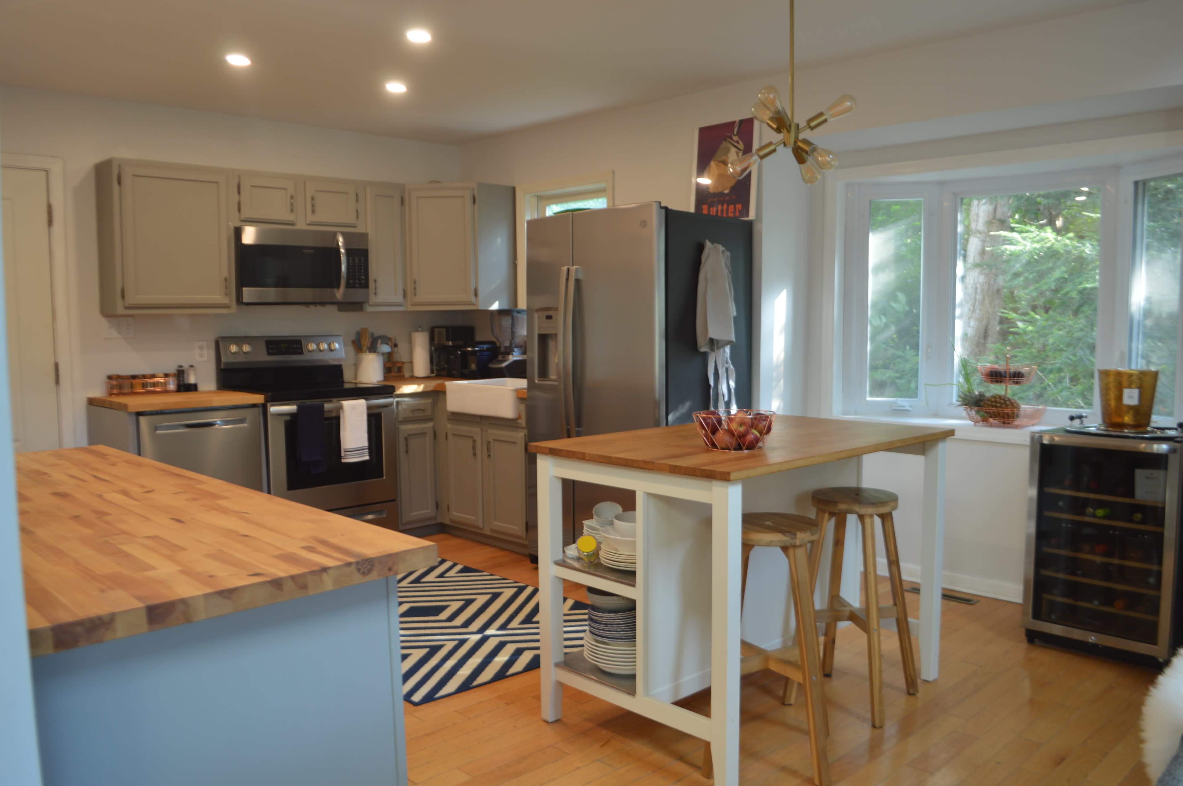 A modern kitchen features grey cabinets, stainless steel appliances, a wooden island with seating, and bright natural light from a nearby window.